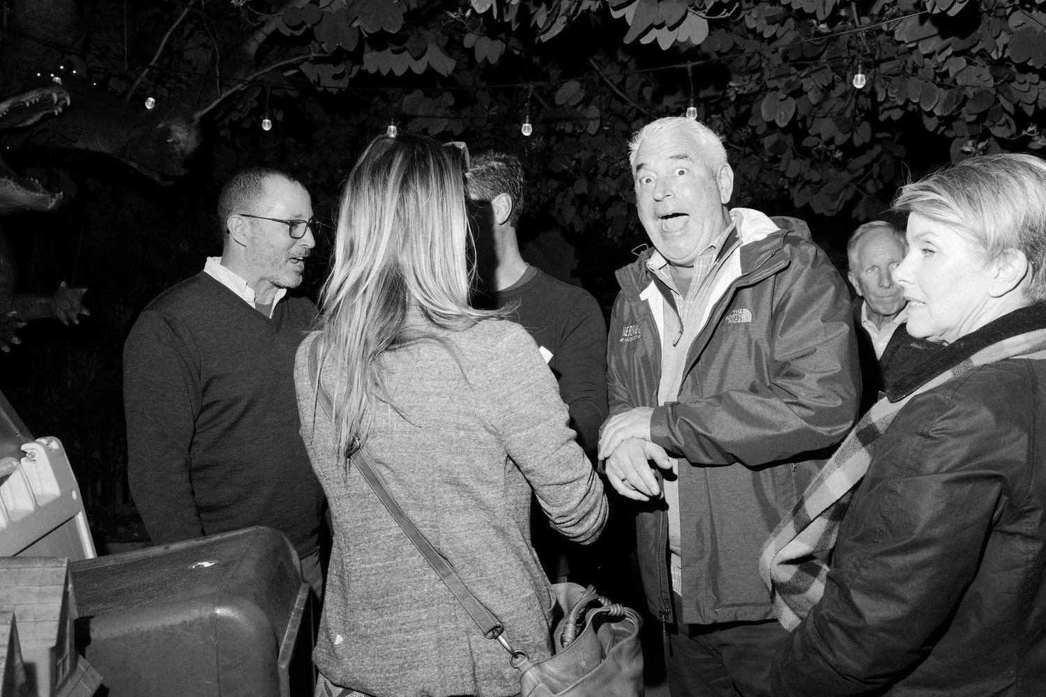 A candid black and white photo of people engaged in lively conversation at an outdoor gathering.