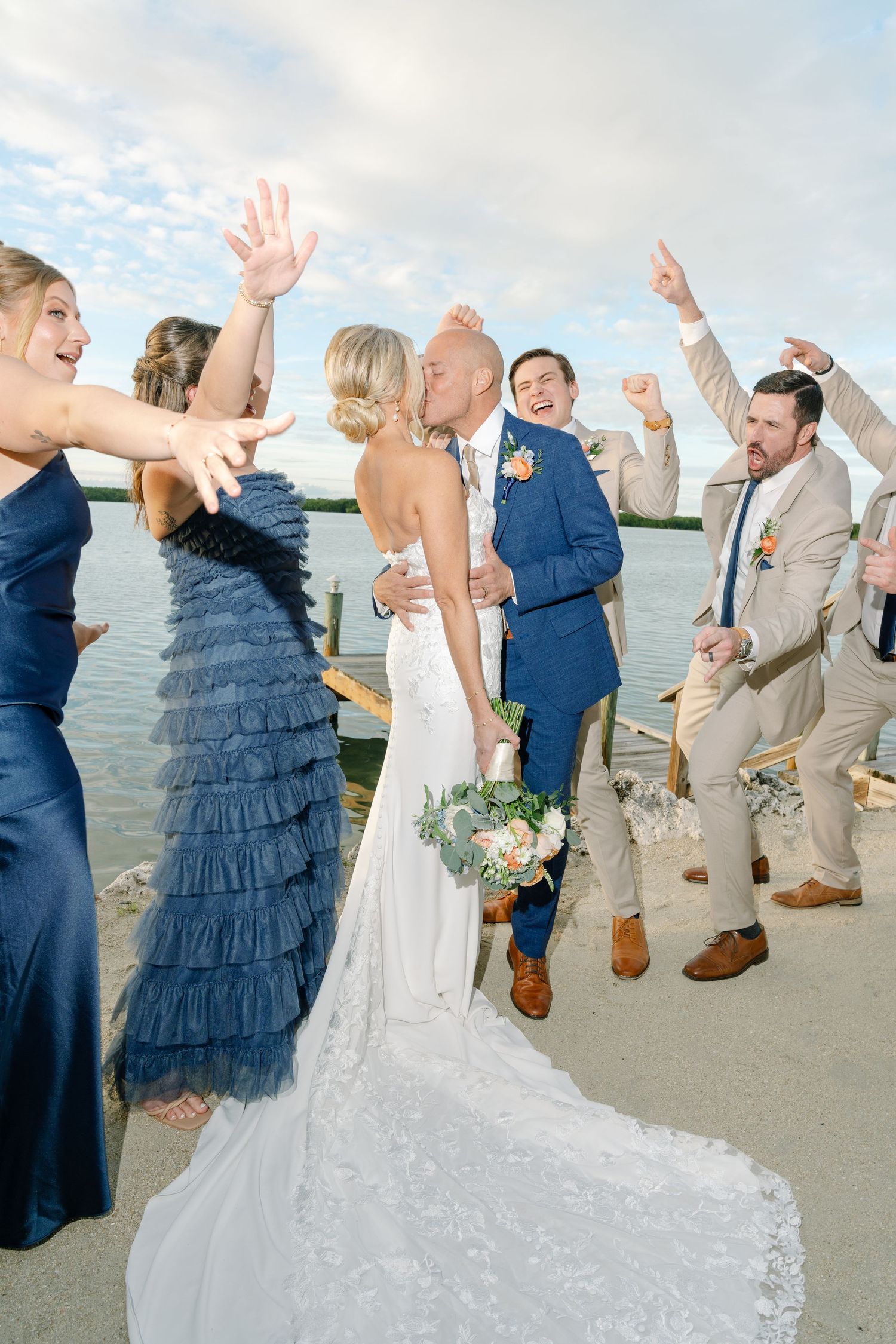 Wedding party celebrates joyfully on a beach with ocean views at sunset.