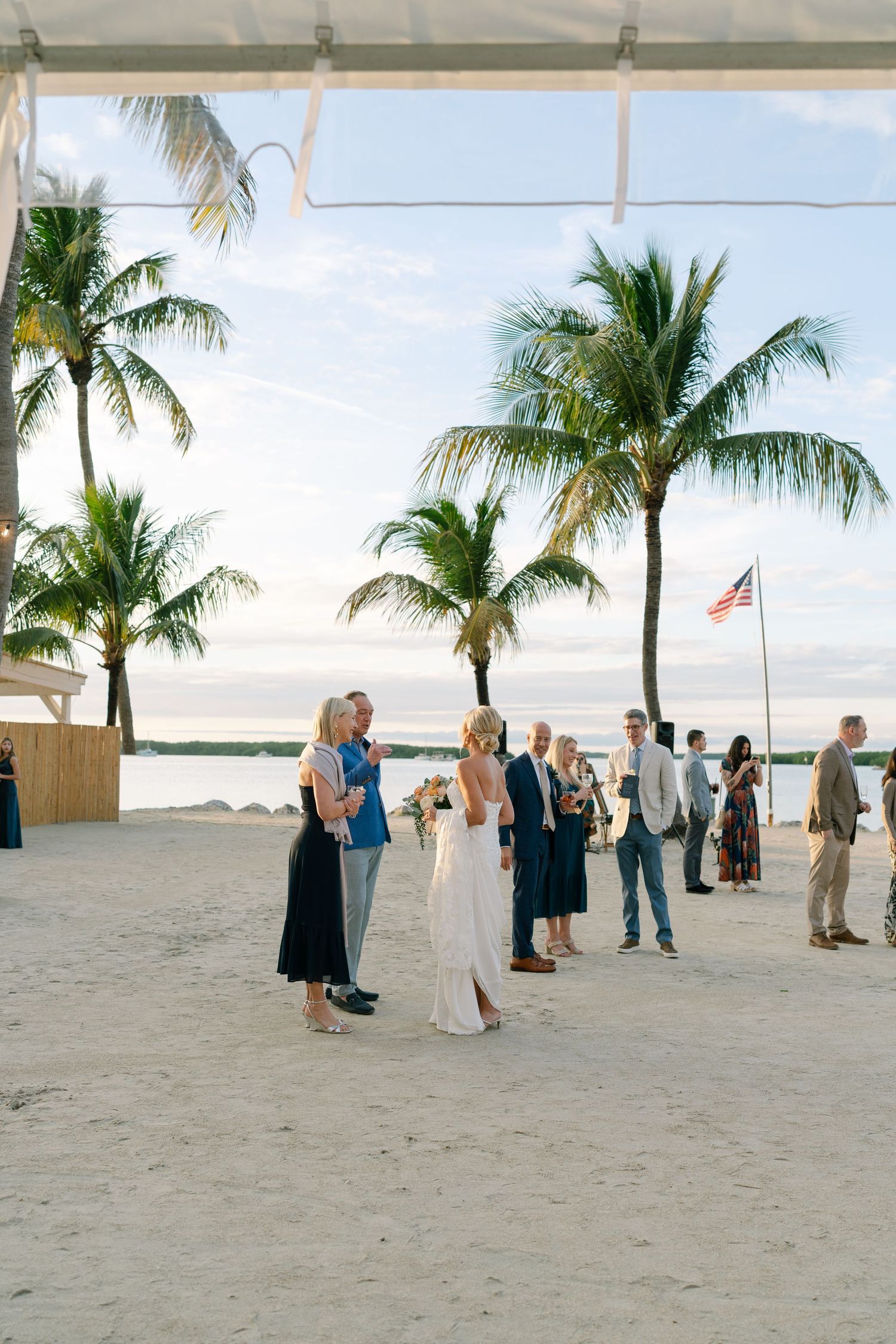 Wedding guests gather on a beachfront patio with palm trees and an American flag in the background.