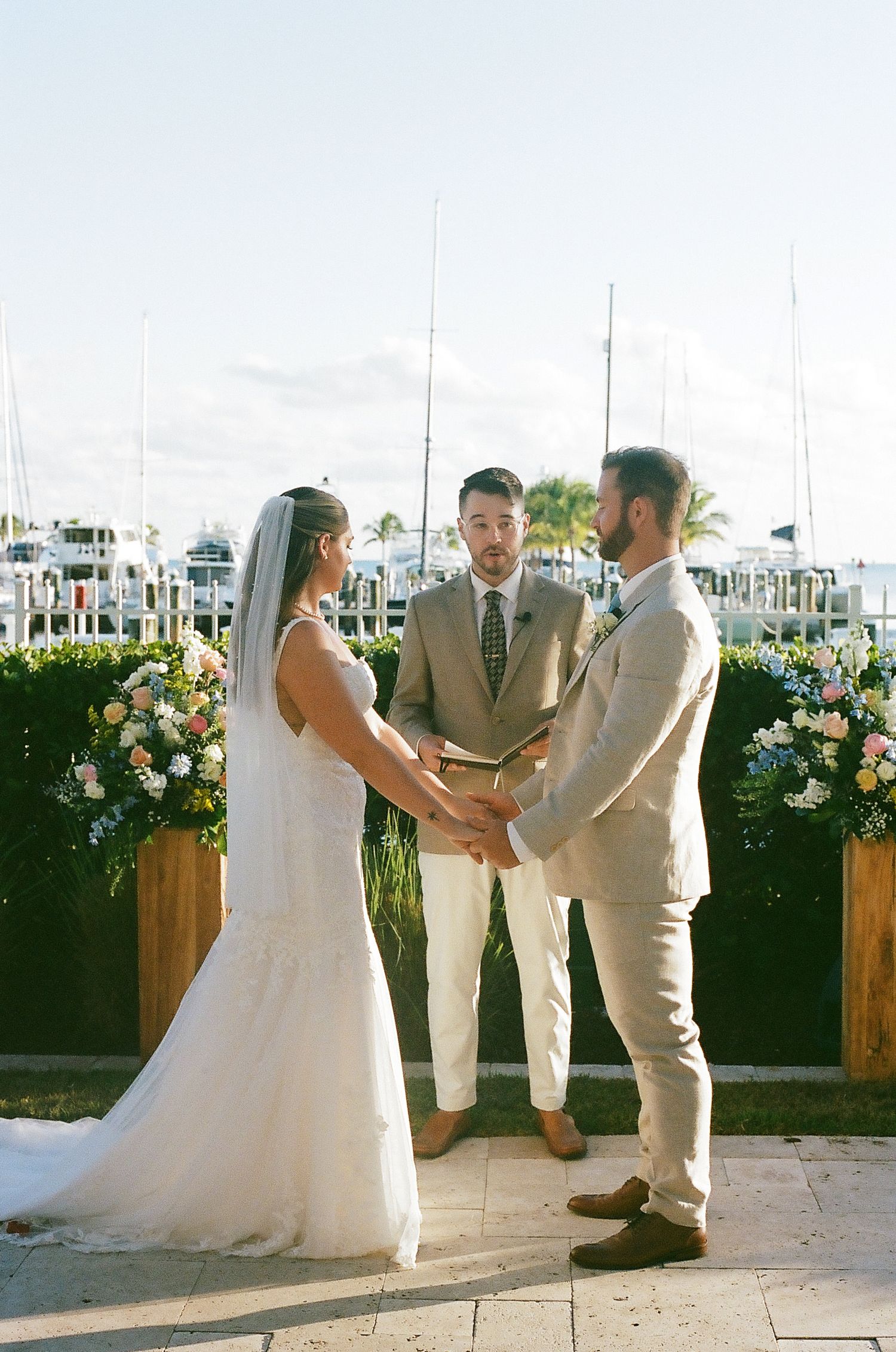 A wedding ceremony takes place at a marina with floral arrangements and boats in the background.