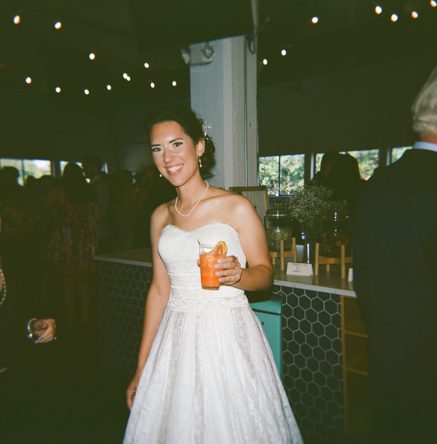 A person in a flowing white dress holds a drink while posing in an indoor setting with twinkling lights in the background.