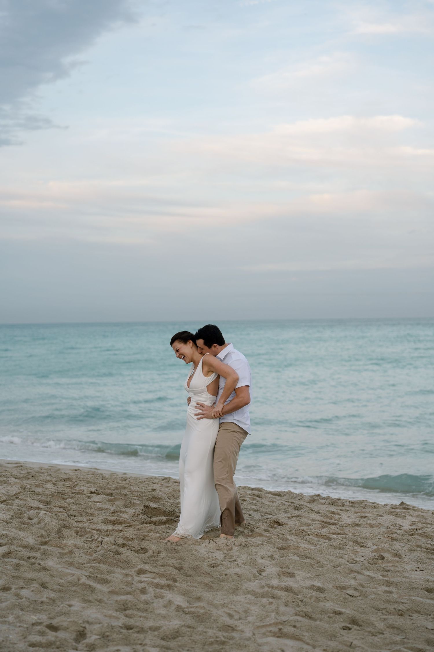 Couple in white and khaki embracing on sandy beach with gentle waves in background.