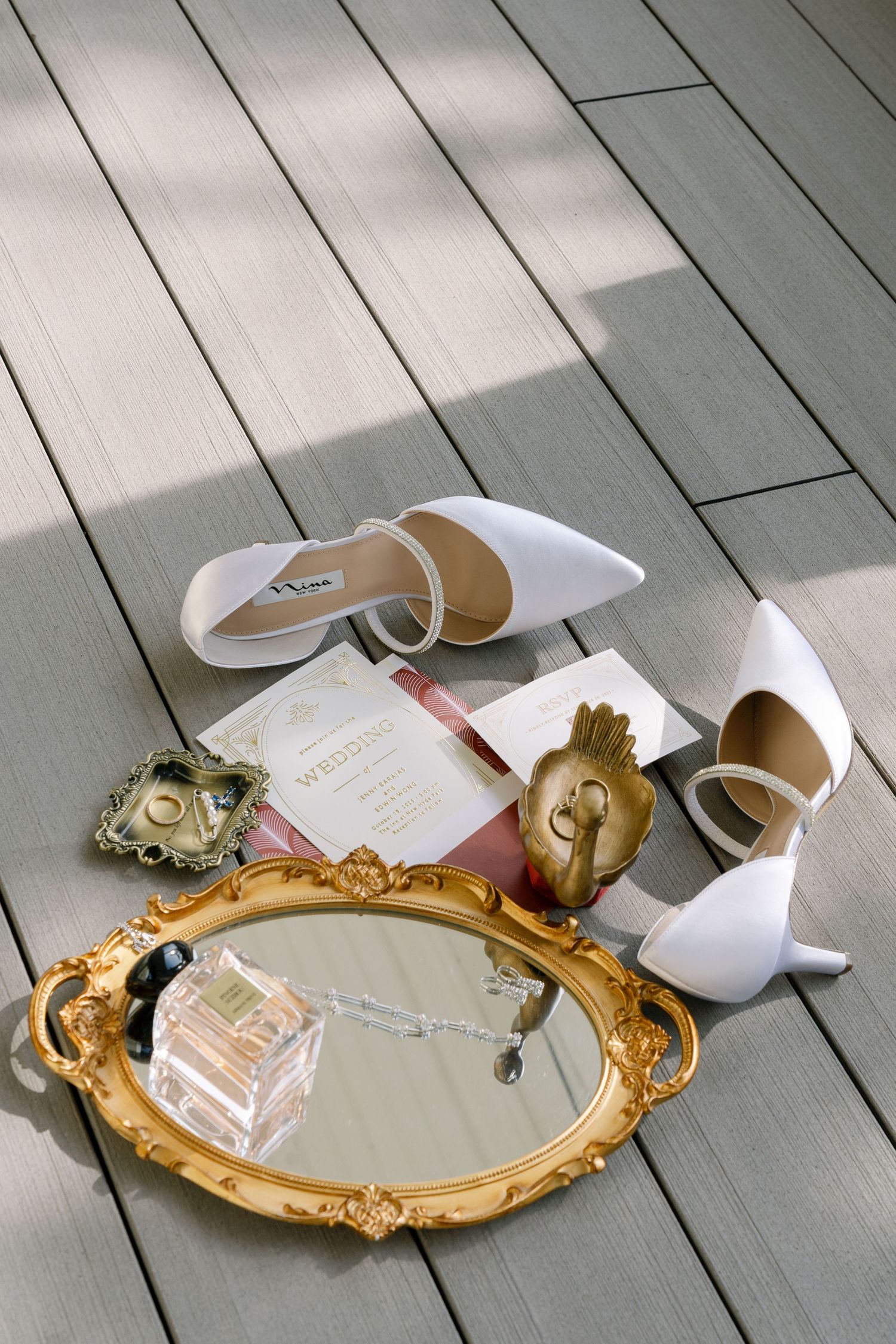 Wedding accessories including a vintage gold mirror, white shoes, and invitation cards arranged on wooden decking.