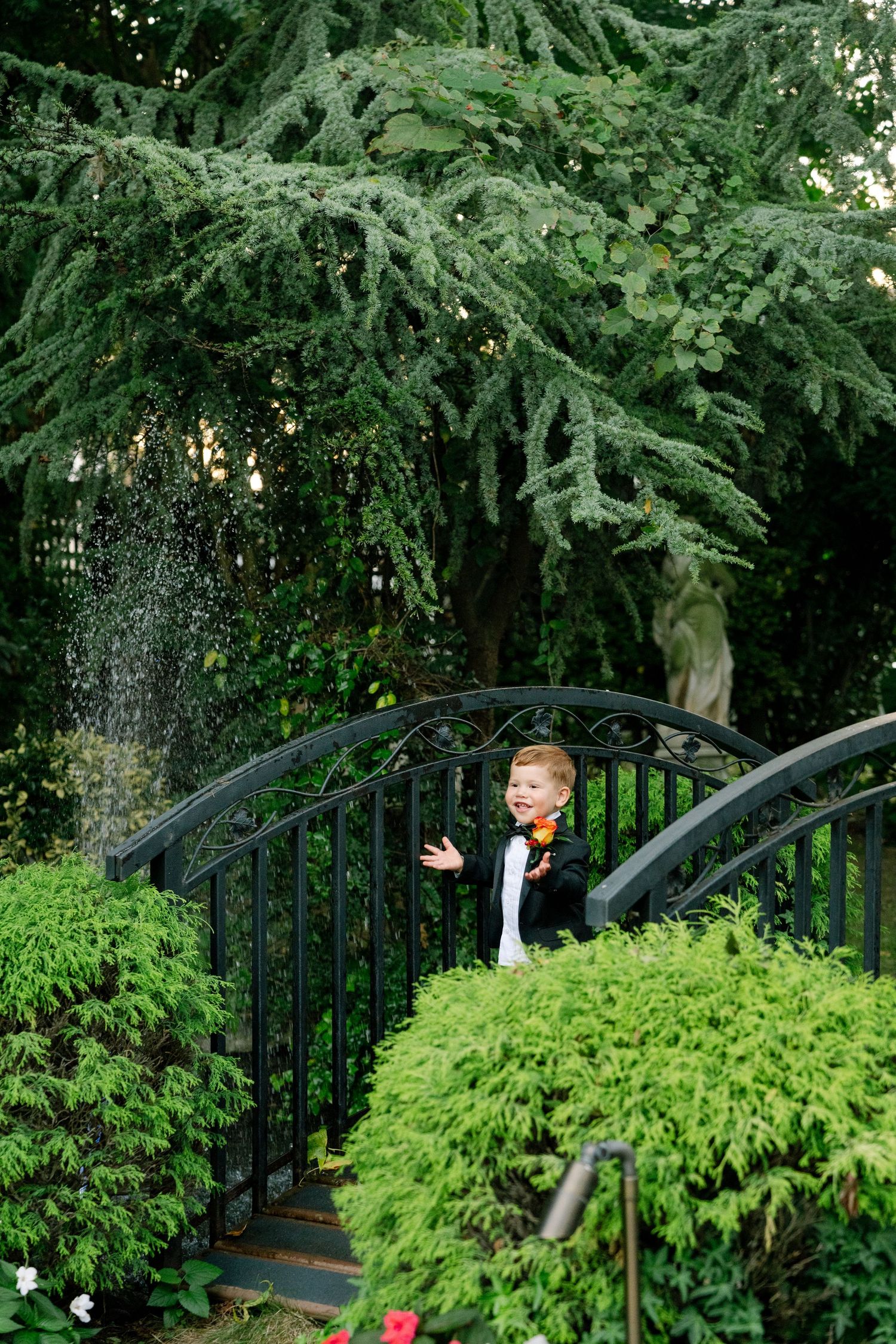 Child in formal wear playing near ornate black garden gate surrounded by evergreens.