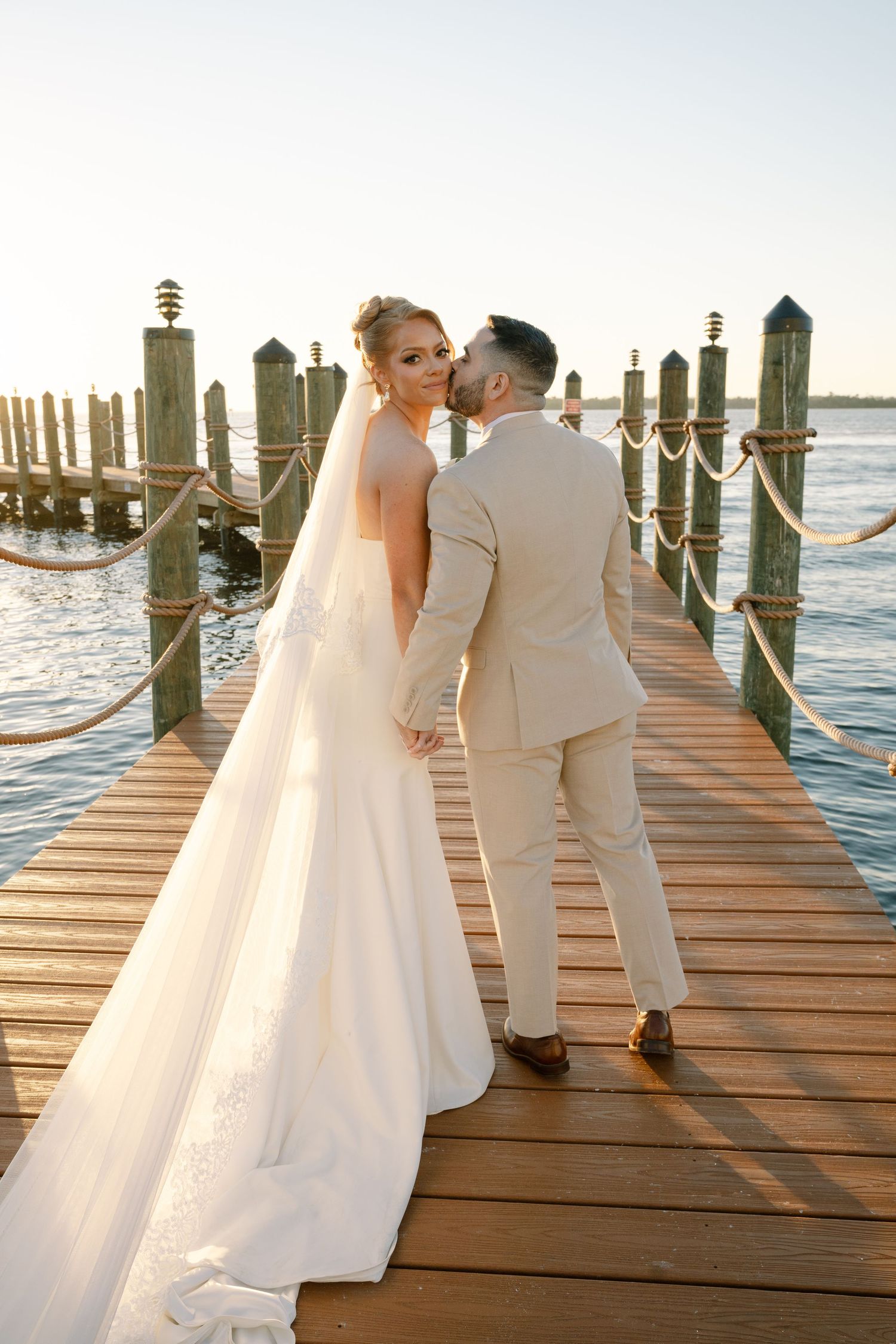 A couple in wedding attire shares an intimate moment on a wooden pier at sunset by the ocean.