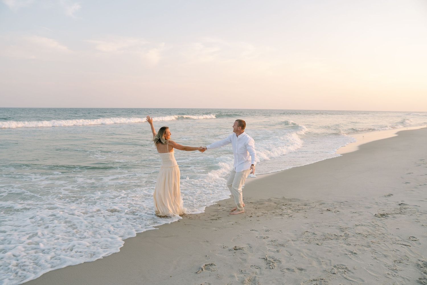 A couple in white outfits holds hands while dancing together on a peaceful beach at sunset.