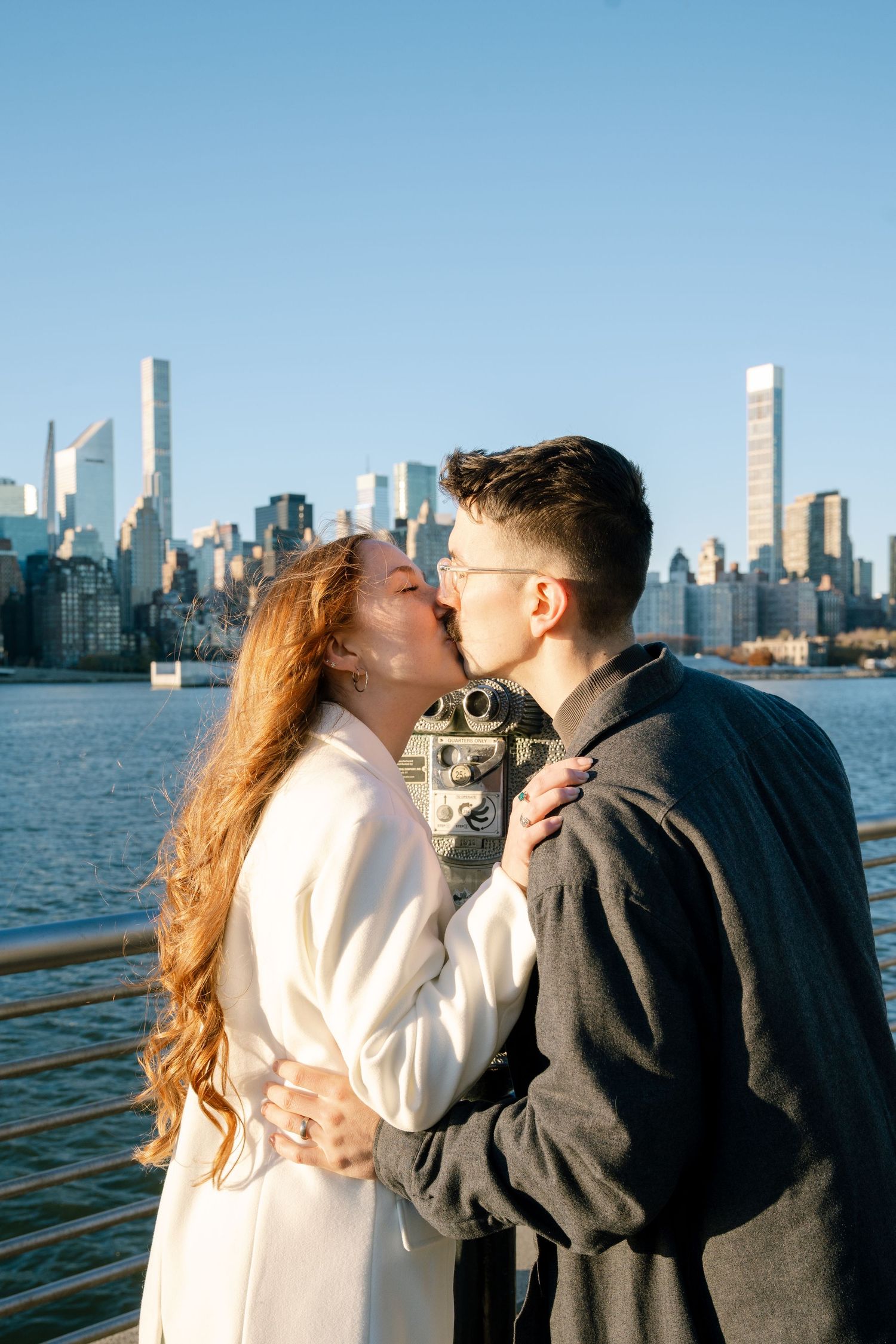 Romantic couple embracing with New York City skyline in background during golden hour.