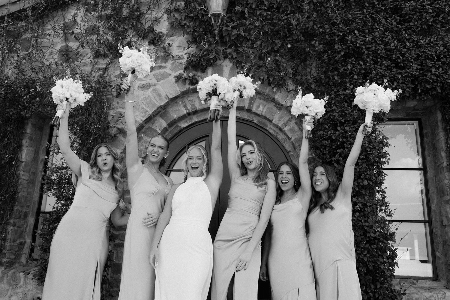 Wedding party in white dresses poses together holding bouquets of white flowers in an elegant garden setting.