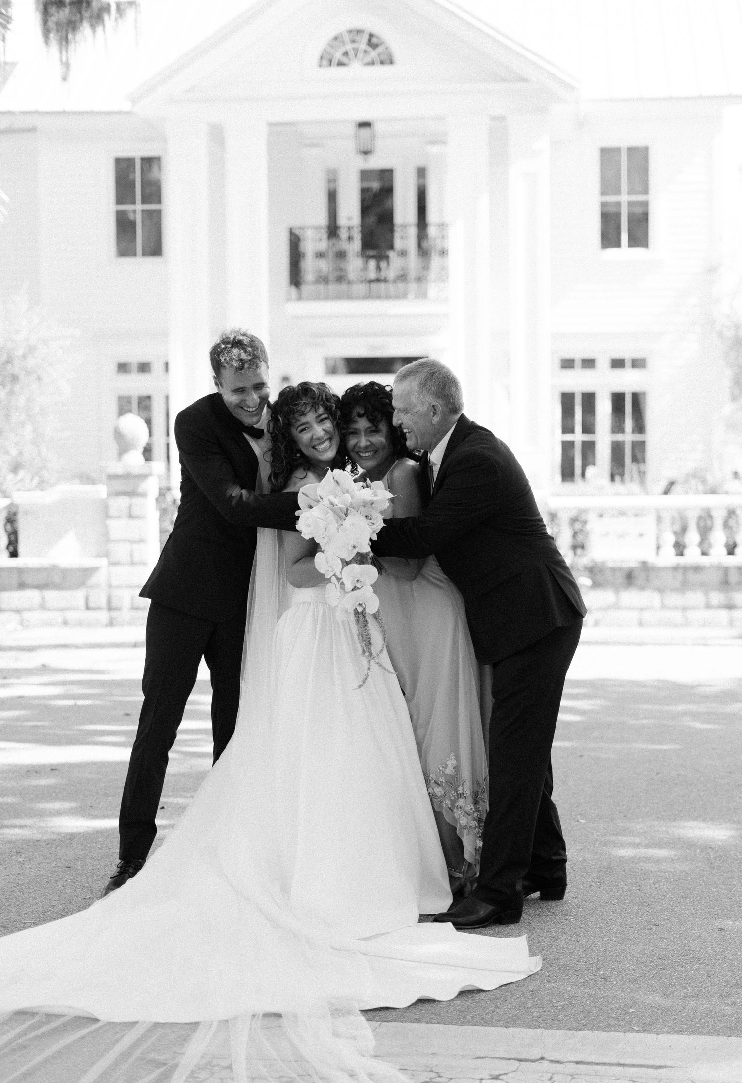 A black and white photo capturing a touching wedding moment with the bride in a flowing white gown outside a grand mansion.