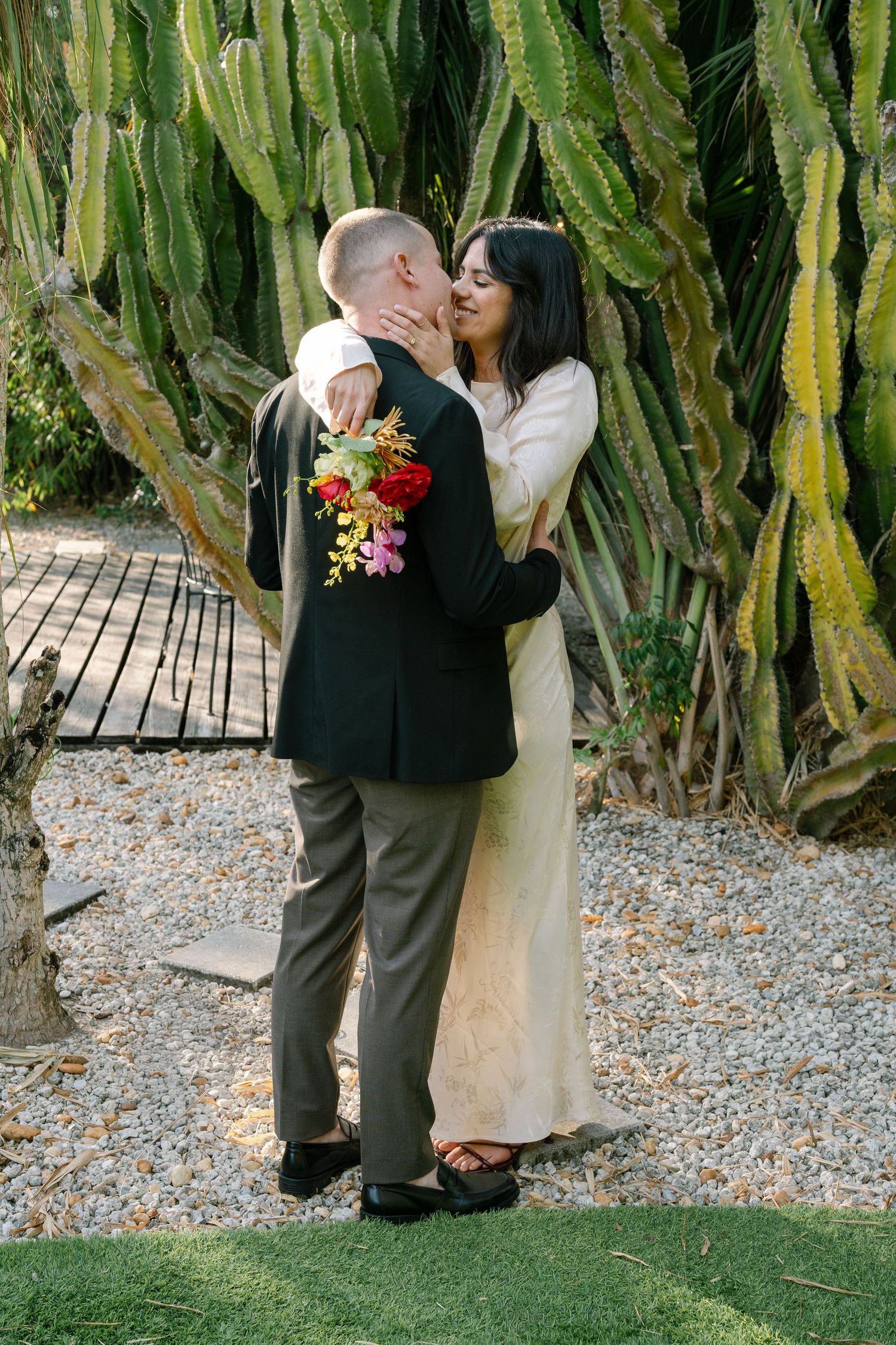 A couple stands together on a gravel path surrounded by tall desert plants.
