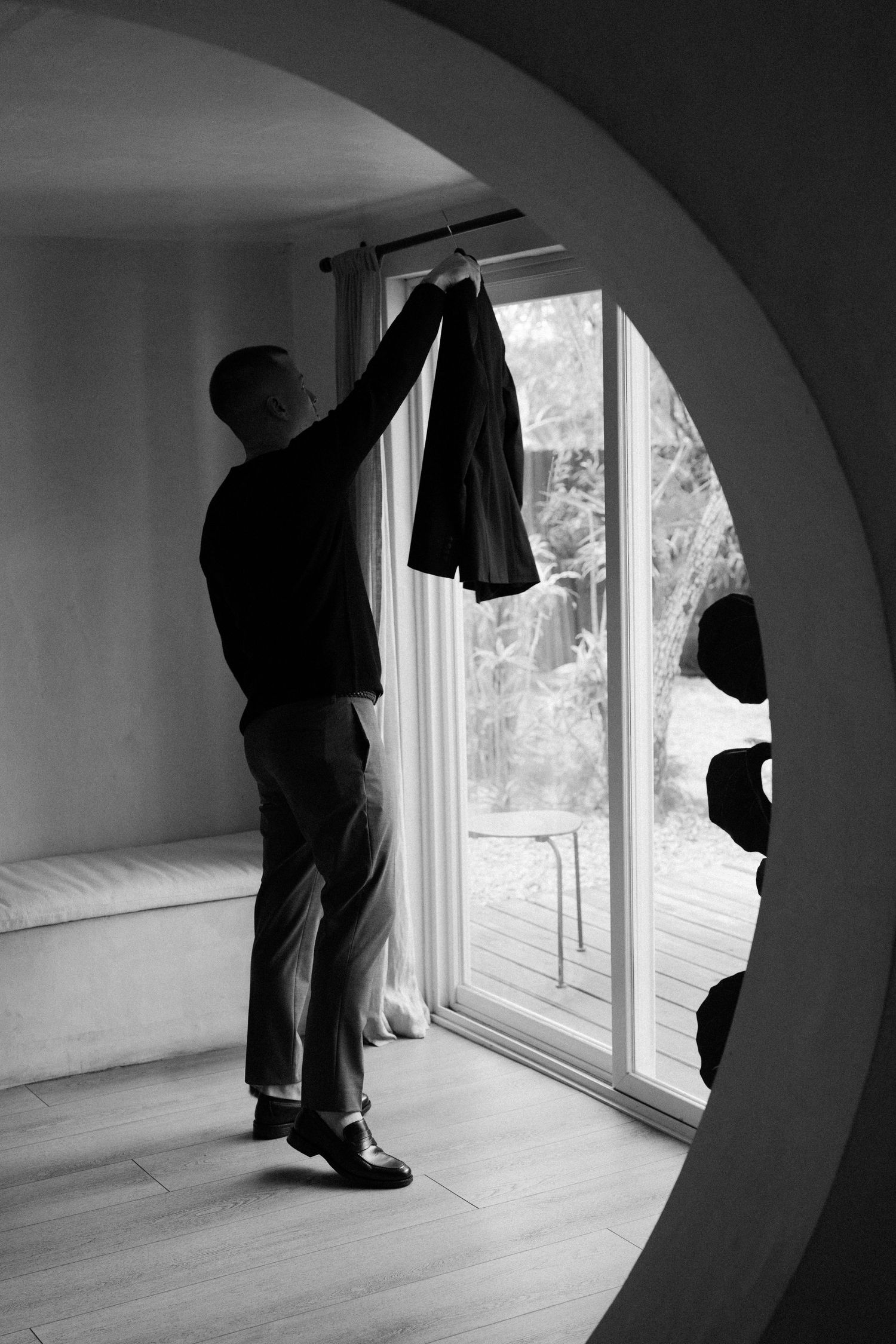 A silhouetted figure hangs a jacket by a large arched window in a minimalist interior space.