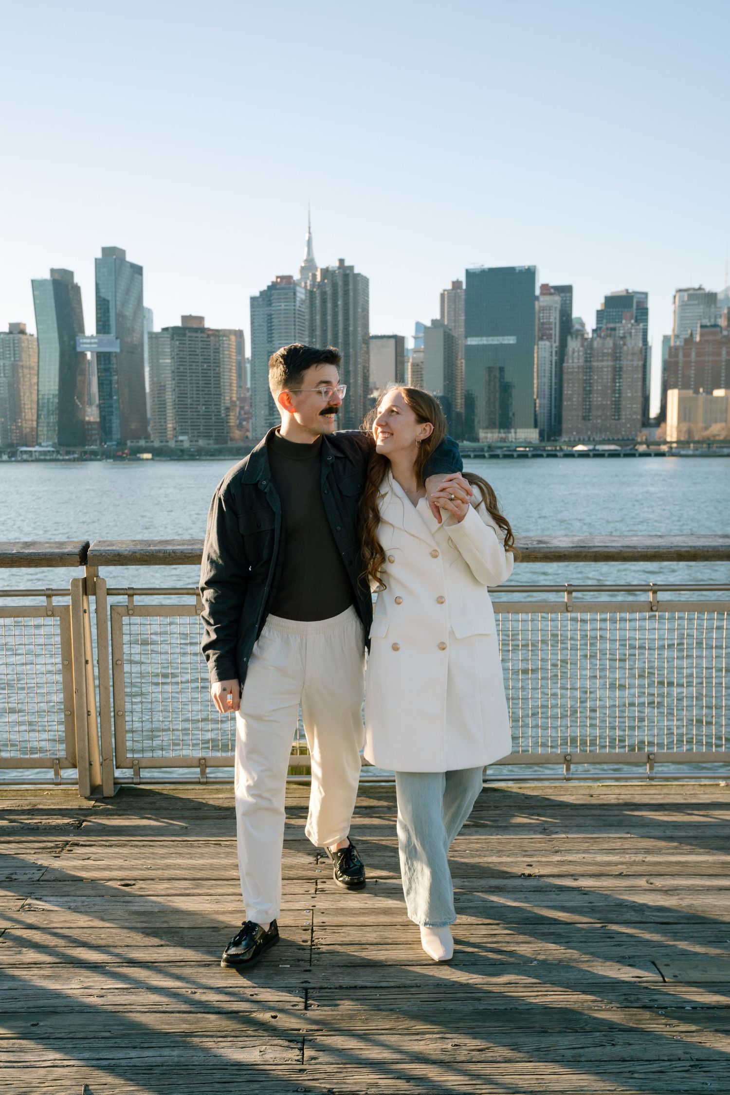 A couple shares playful moments on a pier with Manhattan's skyline visible across the water.
