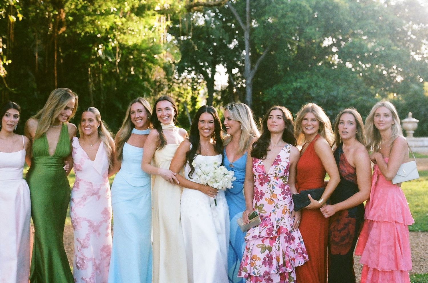 Group of women in colorful formal dresses smiling together outdoors at a wedding celebration.