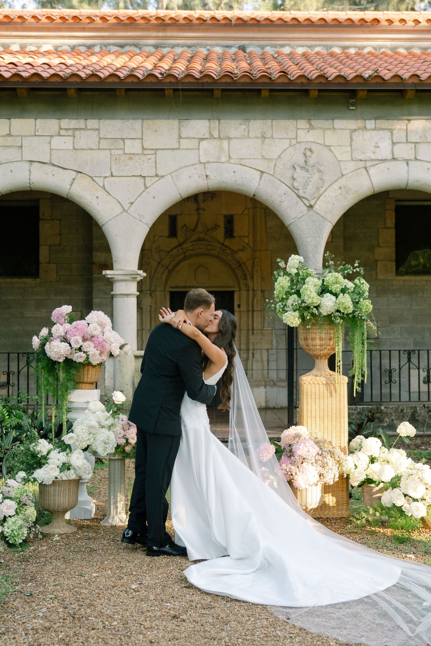 Bride and groom share first kiss outdoors at stone chapel venue surrounded by elegant floral arrangements of pink and white blooms.