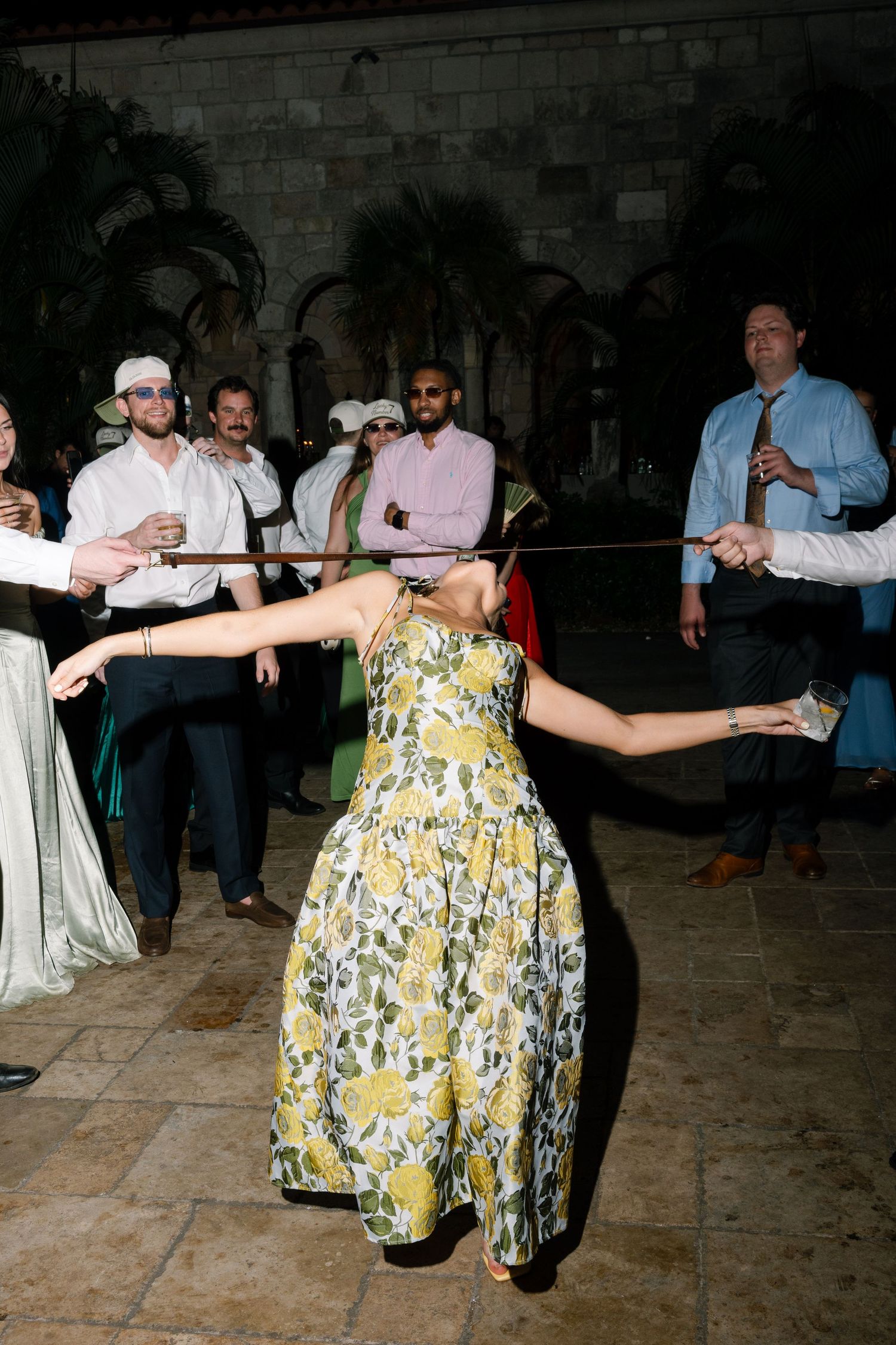 Wedding guest in floral dress doing the limbo under a stick surrounded by cheering guests at reception.