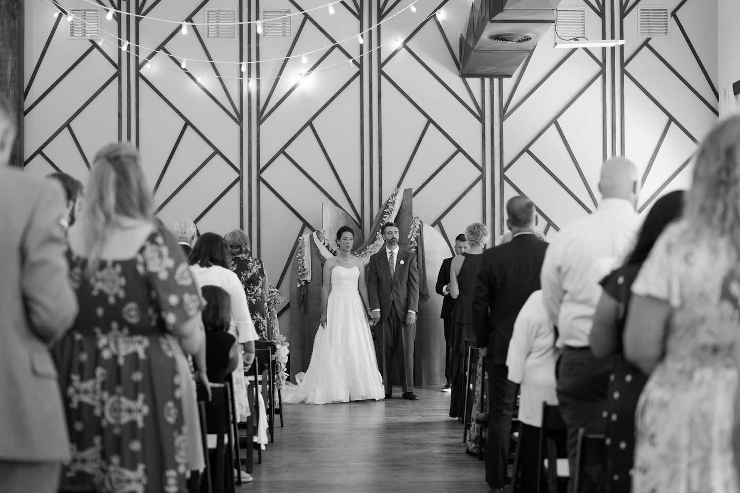 Wedding ceremony in progress with art deco geometric pattern backdrop and seated guests.