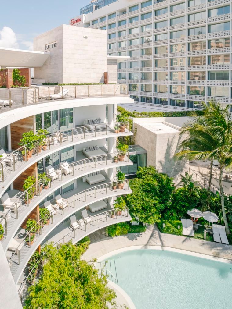 Modern hotel balconies curve around a tranquil turquoise pool surrounded by lush tropical landscaping and palm trees.