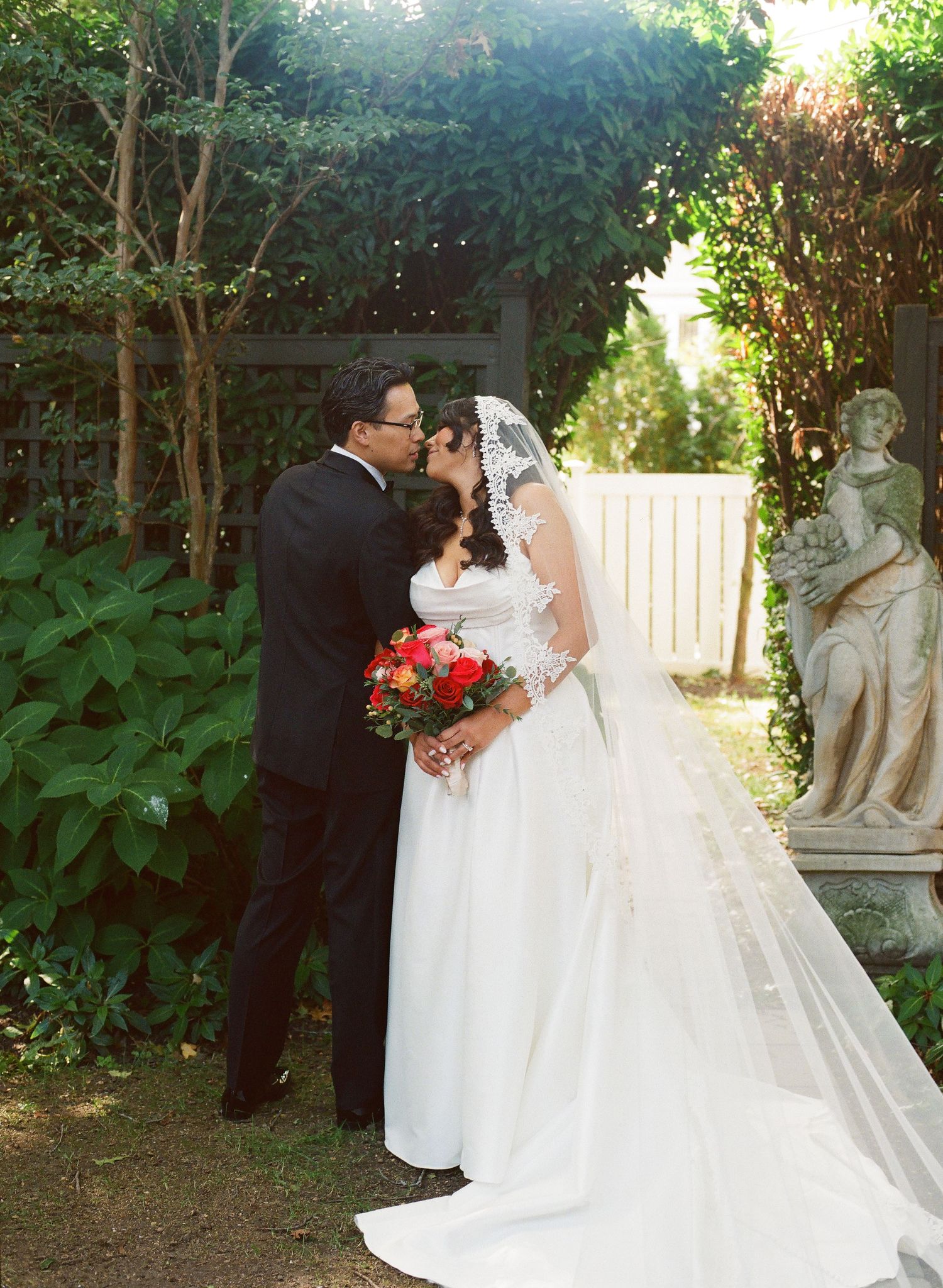 A romantic garden wedding scene with a flowing white dress and veil against lush greenery and decorative statues.