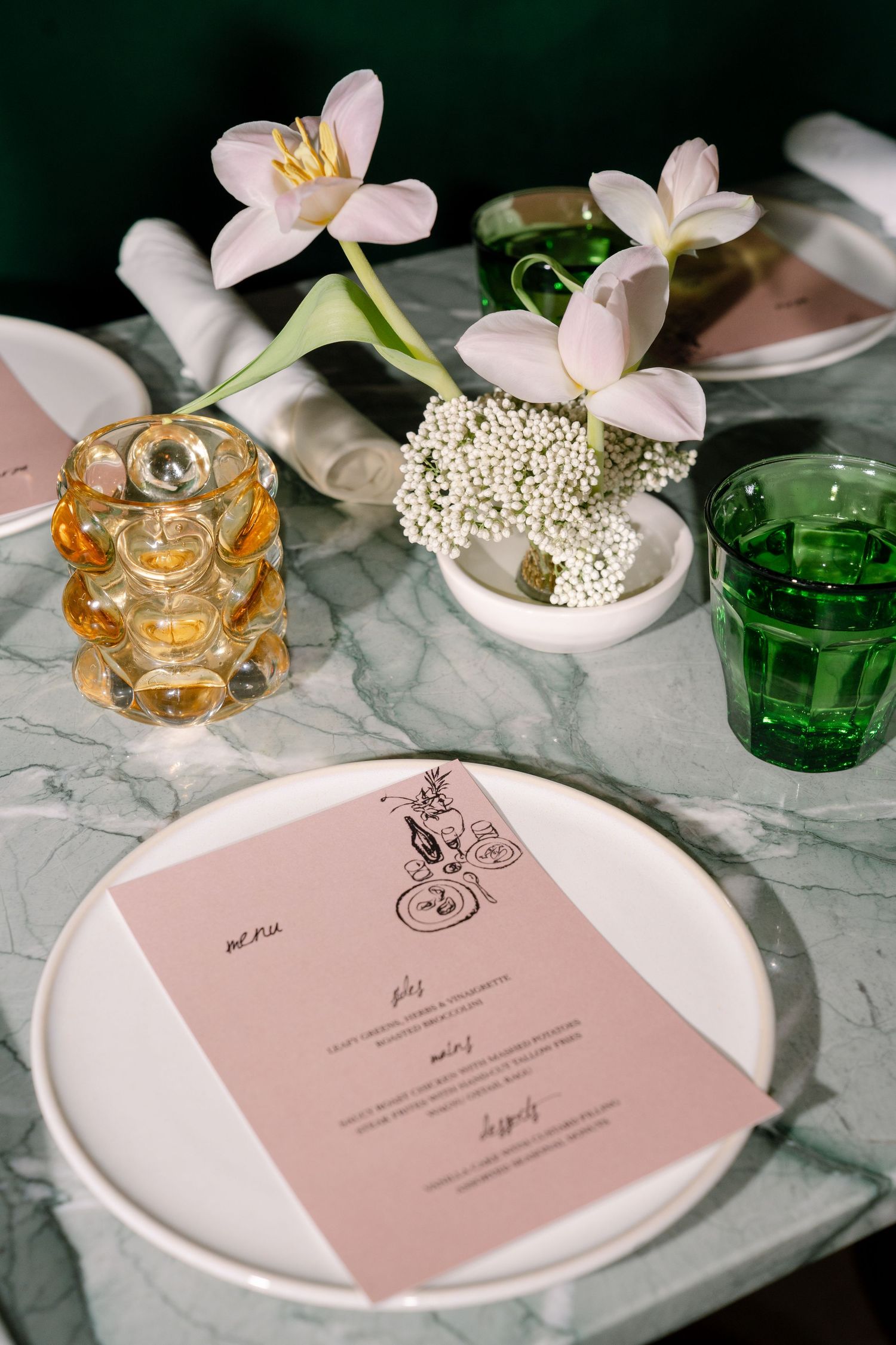 Elegant table setting with pink menus, delicate flowers, and crystal glassware on marble surface.