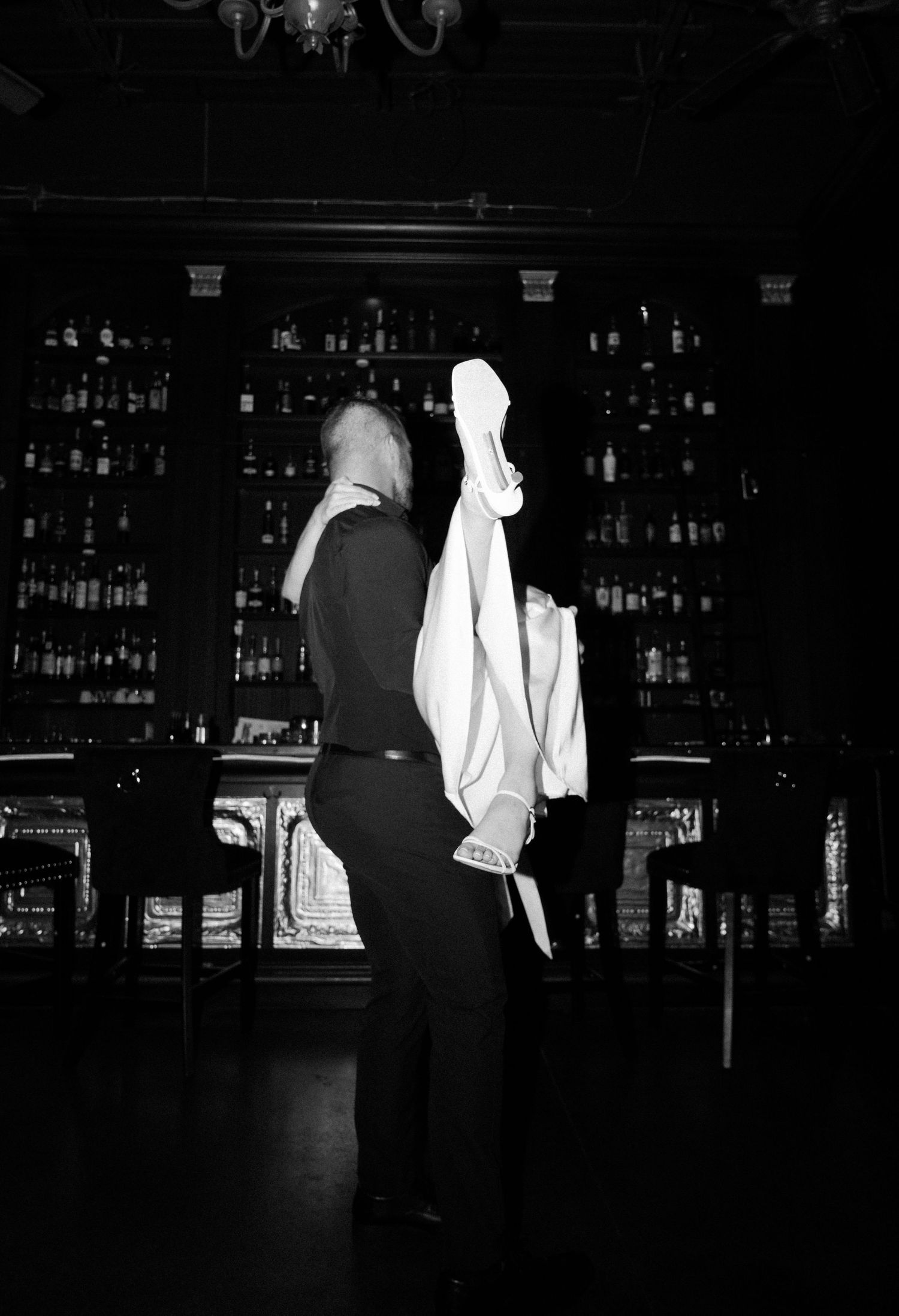 A dark moody black and white photo of silhouettes standing at a bar with bottles illuminated in the background.