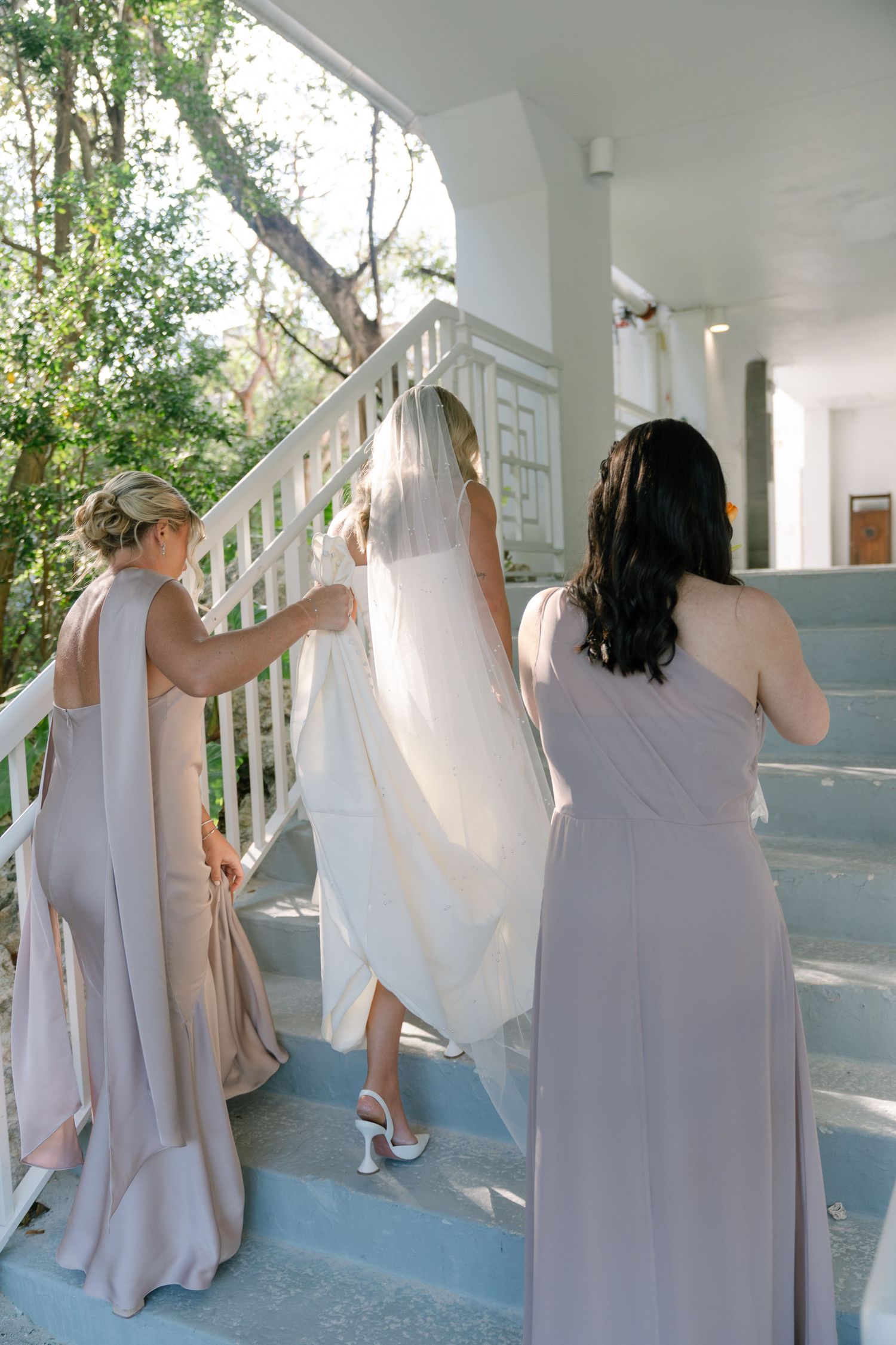 Bridesmaids in flowing taupe dresses line up along a white porch railing.