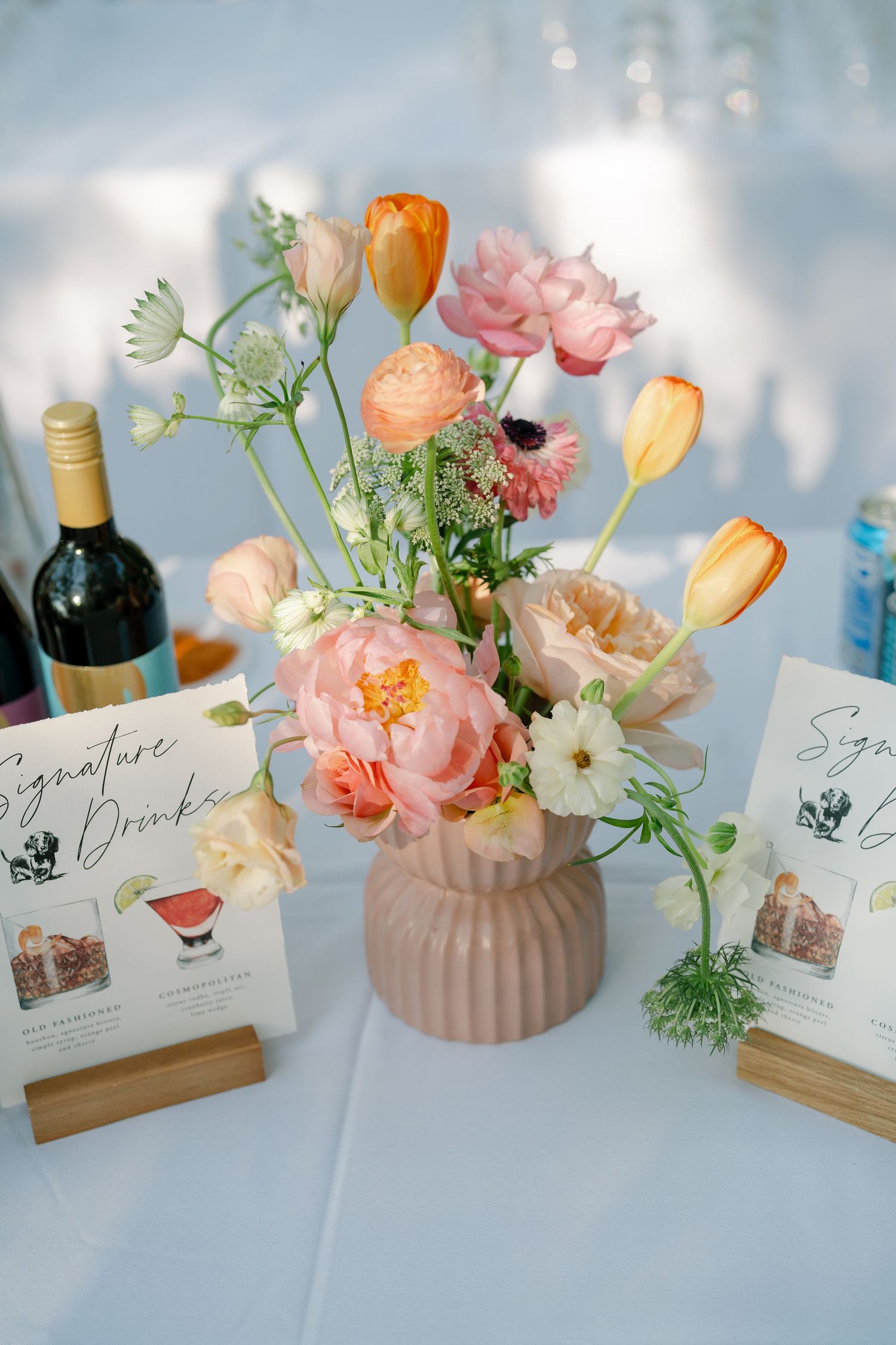 Elegant floral centerpiece with pink and orange blooms in a ribbed vase alongside wine bottle and menu cards on white table.