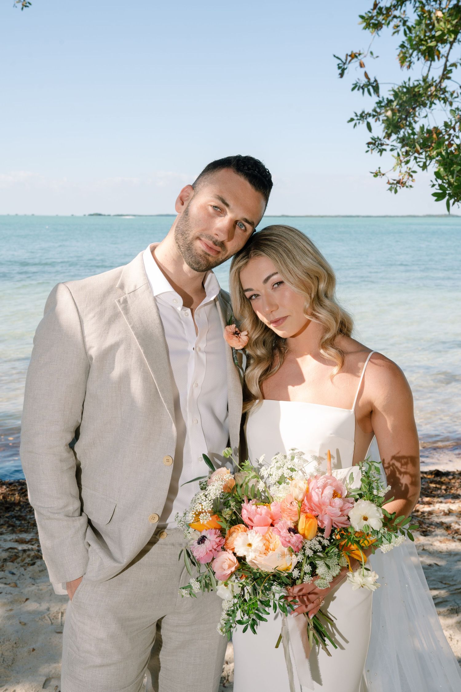 Newlyweds pose for romantic portraits by the ocean with coral and peach floral bouquet.