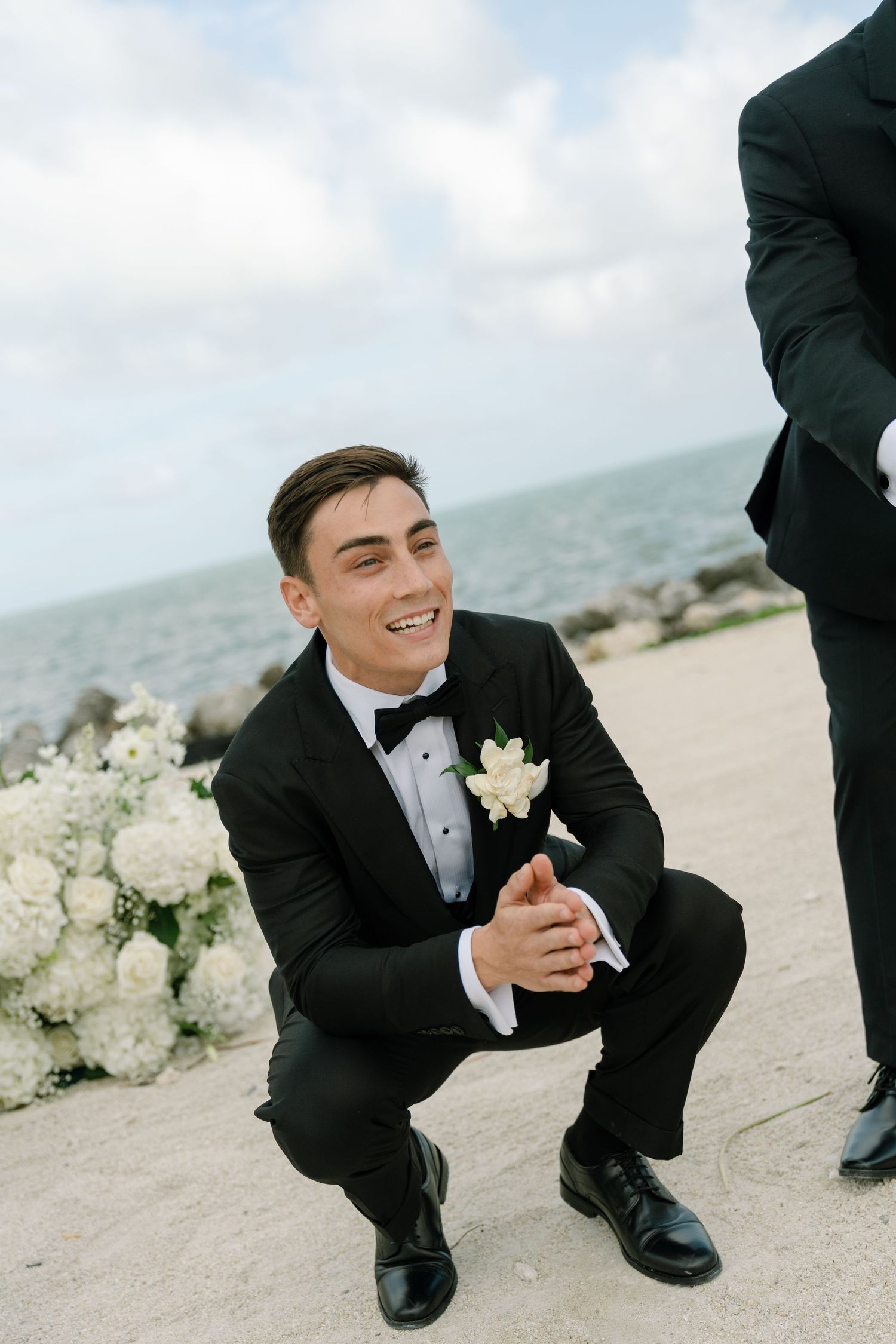 Smiling groom in black tuxedo crouching on sandy beach with ocean and white floral arrangements behind him.