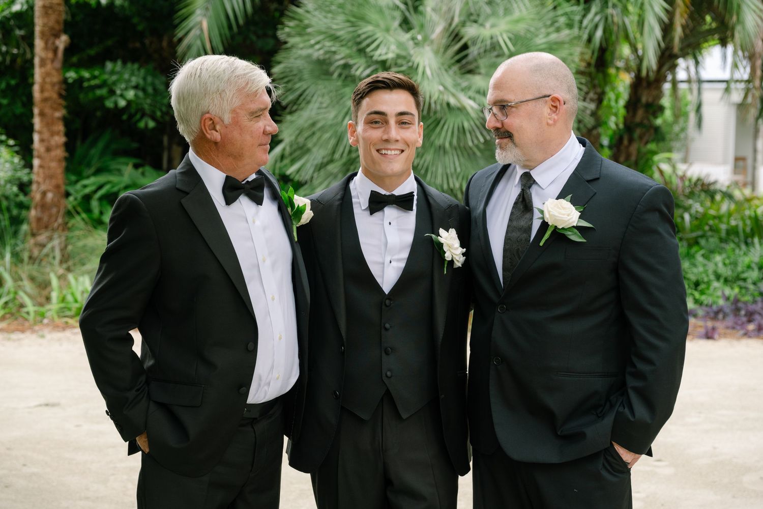 Groom poses with two family members in black tuxedos with white boutonnieres outdoors.
