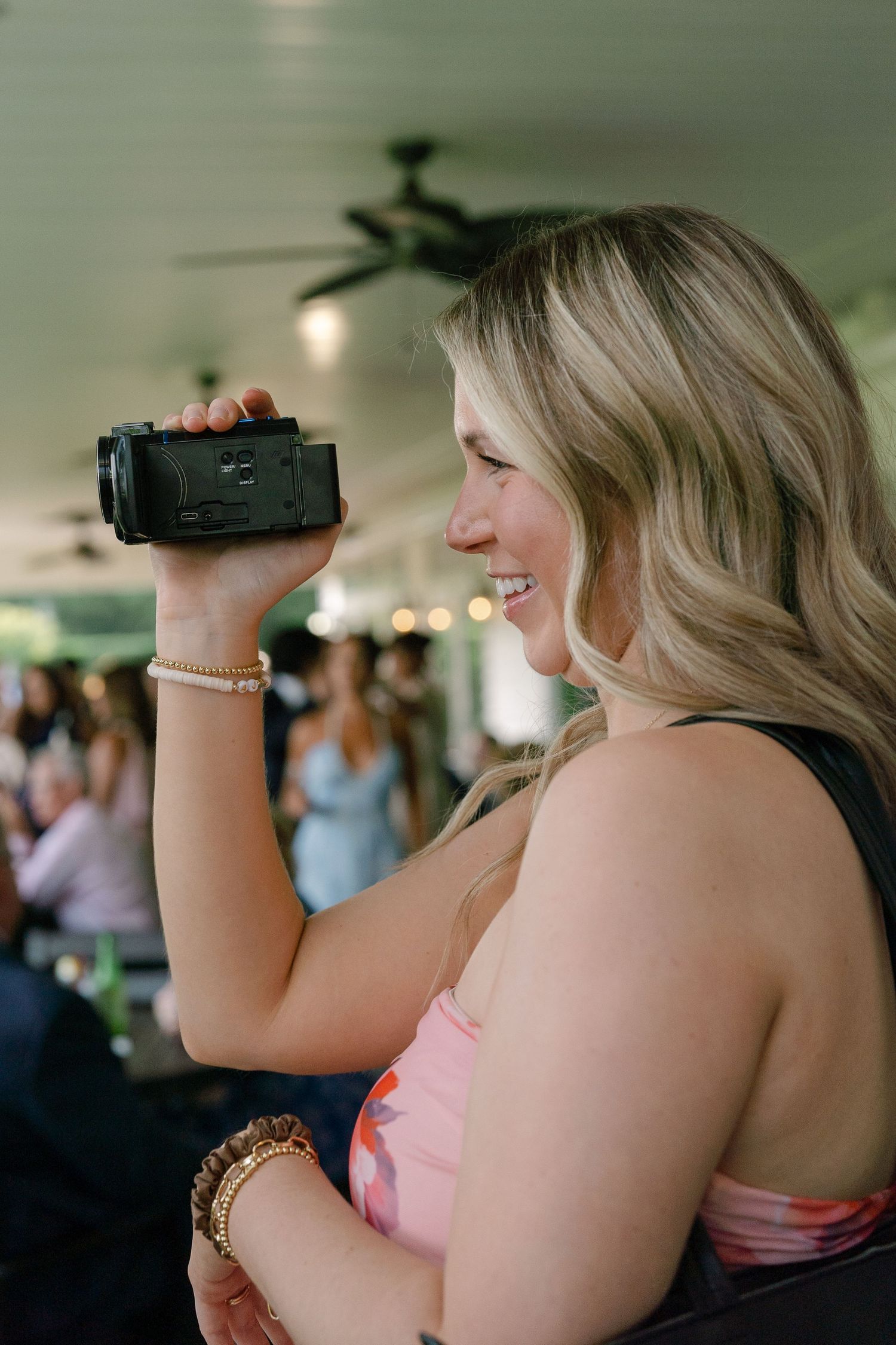 Blonde woman in floral dress smiling while recording wedding reception on handheld video camera.