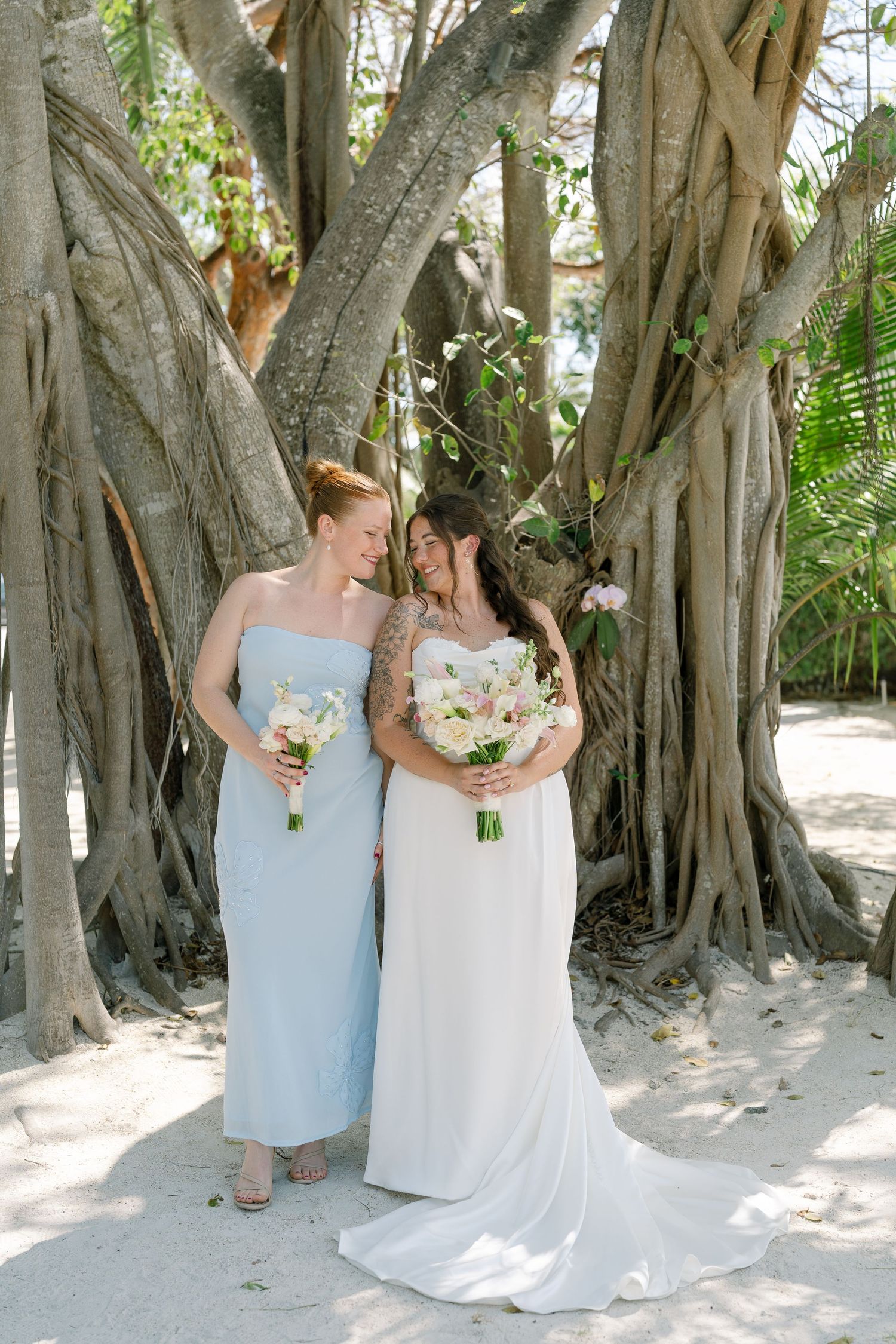 Two brides in white wedding dresses embrace under a large tree on a beach.
