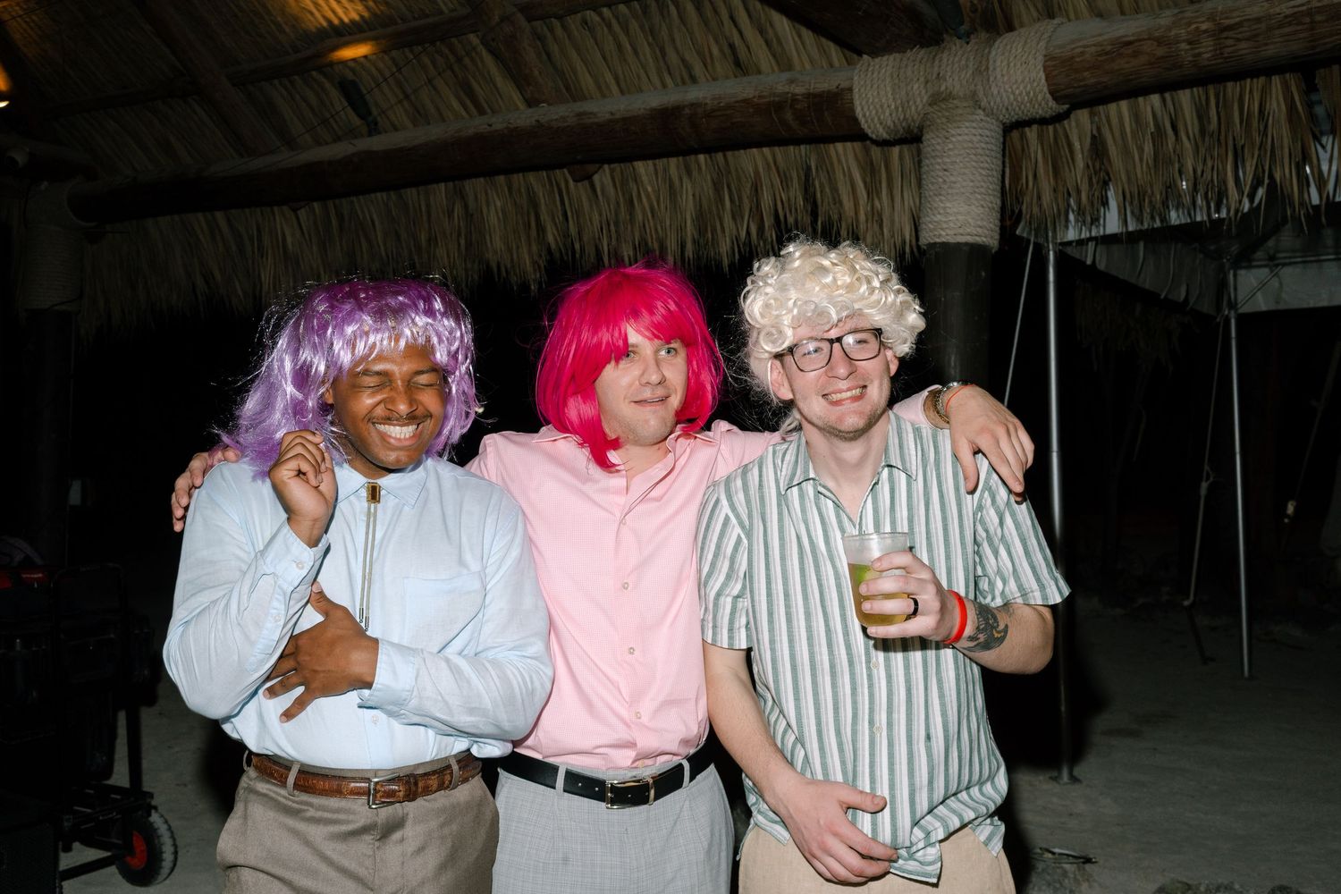 A group of three people wearing colorful wigs and costumes stand together in a rustic setting.