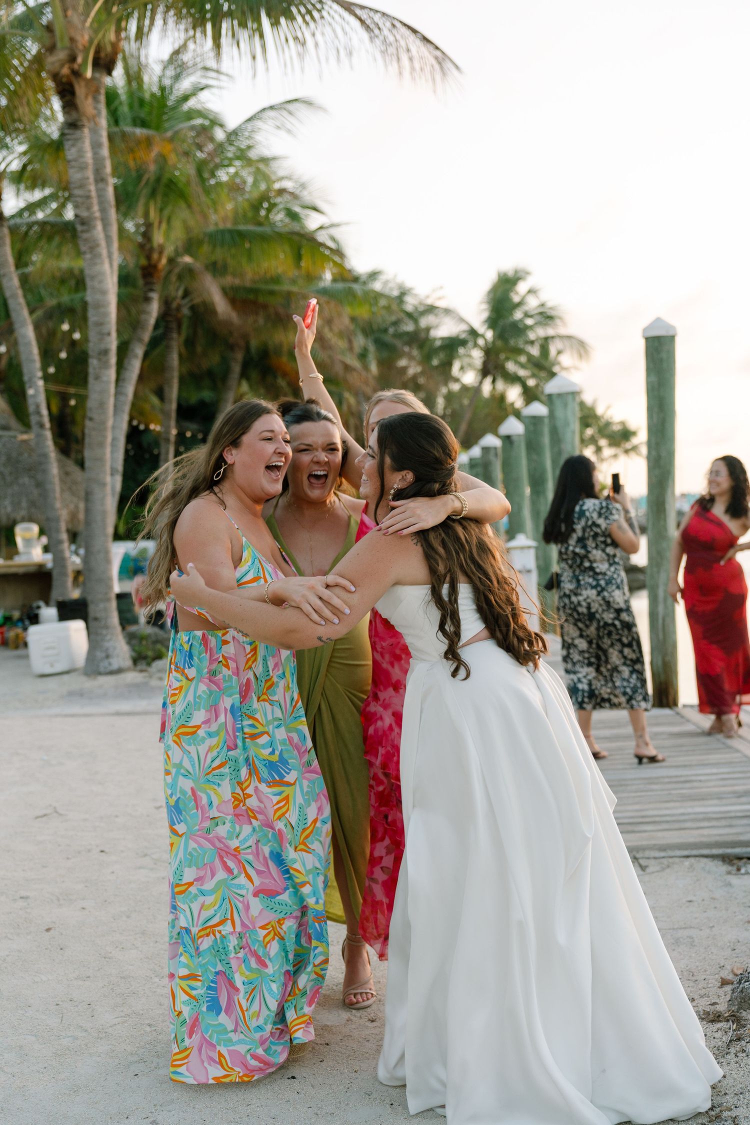 A group of women in colorful dresses hug and embrace on a beach.