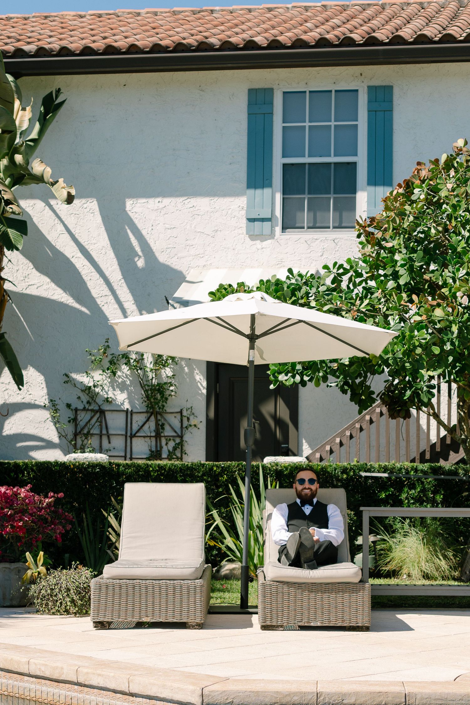 A cozy outdoor seating area with wicker furniture, a white umbrella, and a lush garden surrounding a Mediterranean-style building.