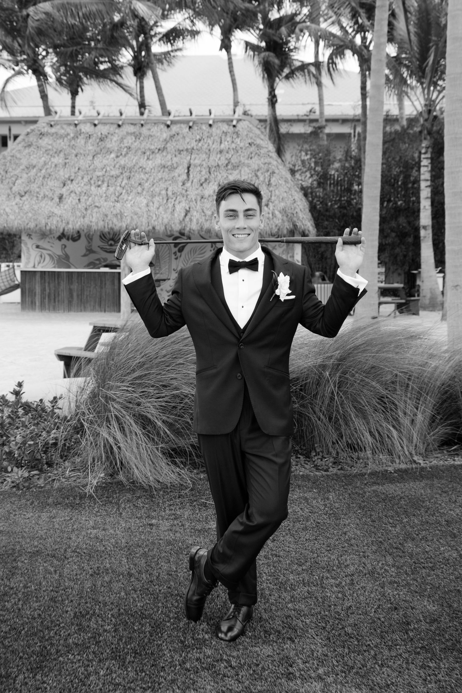 Dapper groom in black tuxedo and bow tie posing playfully outdoors at a tropical resort wedding venue.