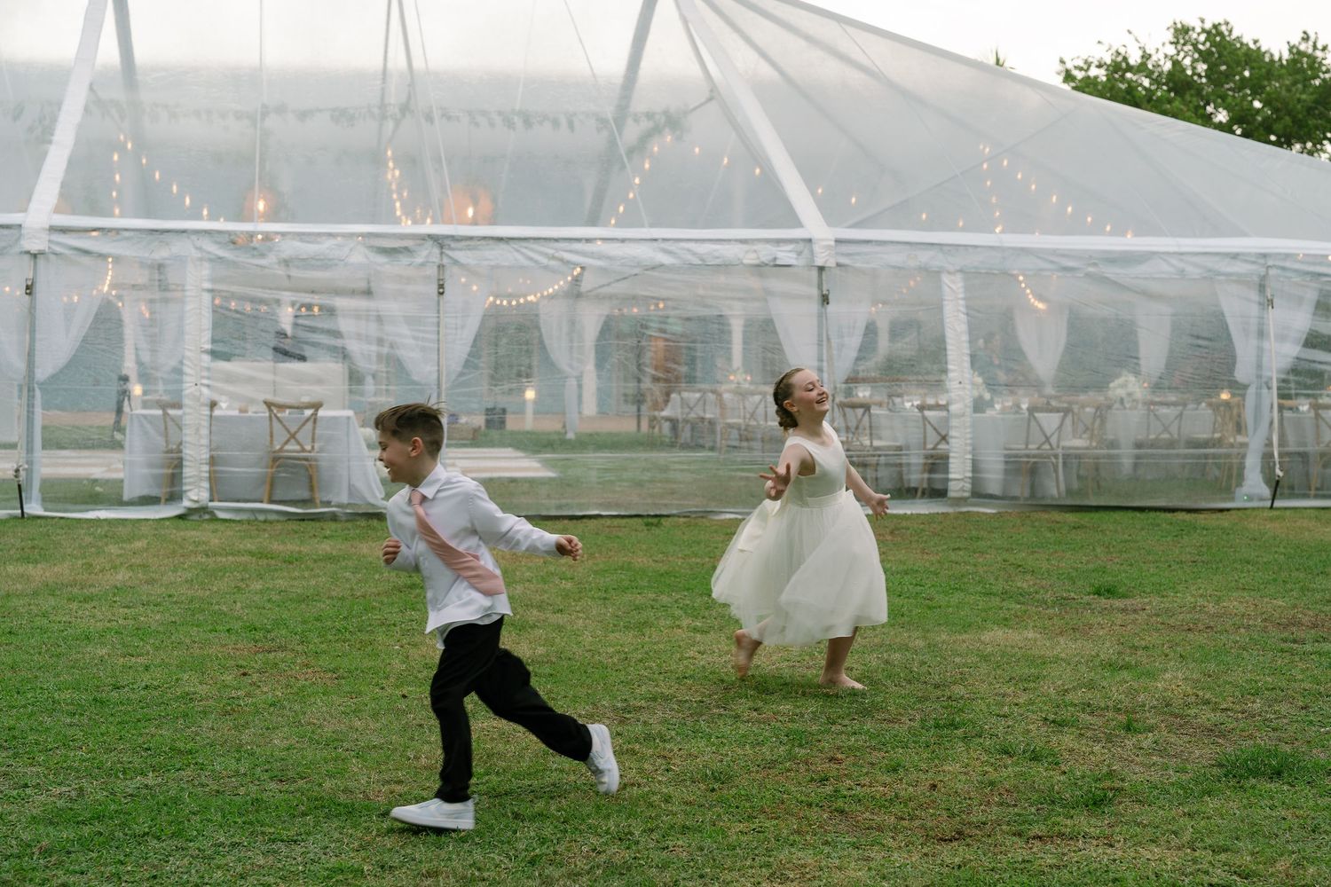 A young boy and flower girl in wedding attire running playfully on the grass outside a clear reception tent.
