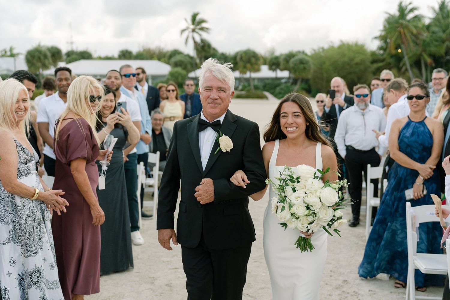 Father walks bride down sandy beach aisle as guests watch outdoor wedding ceremony.