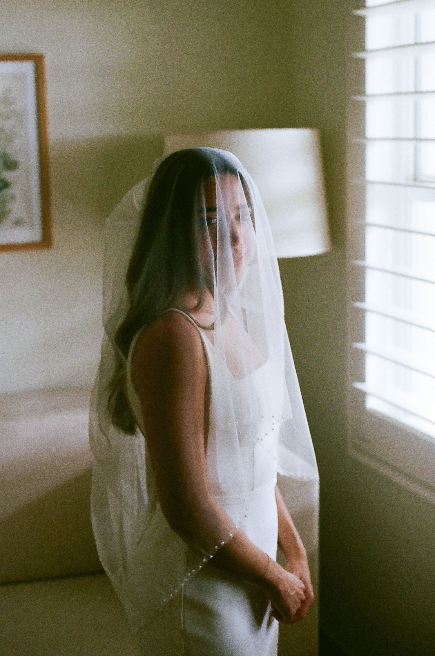 Bride wearing pearl-trimmed veil standing by window in soft warm light indoors.