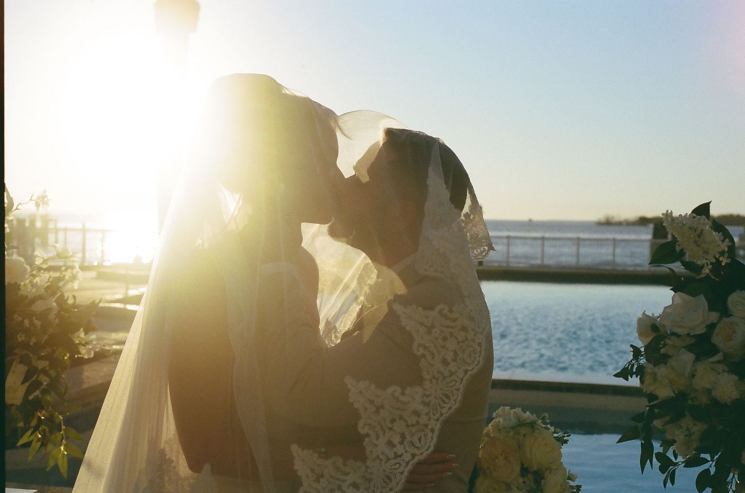 A couple embraces in golden sunlight by a waterfront during sunset with lens flare illuminating the romantic scene.