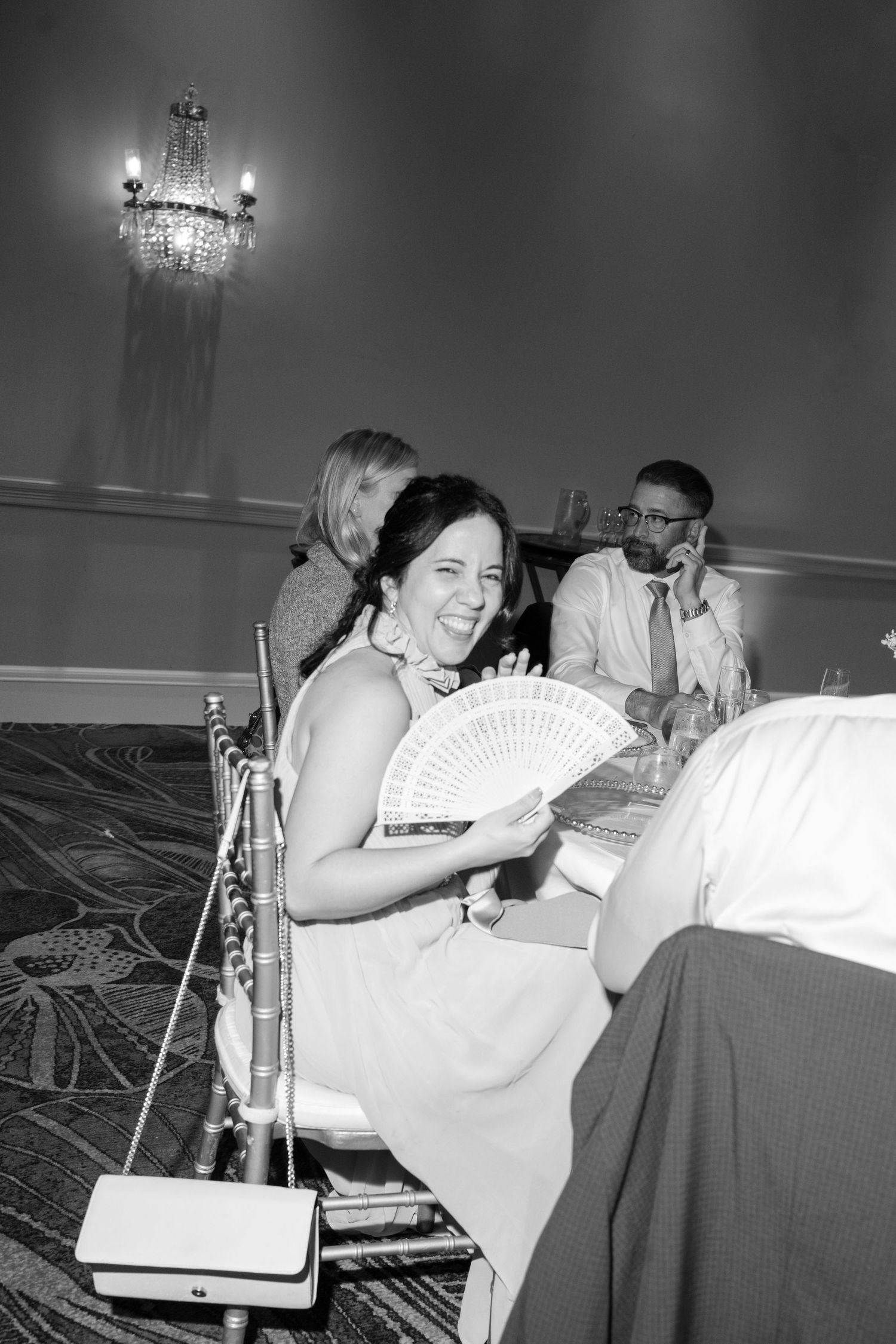 Wedding party members laugh while sitting in chairs holding decorative paper fans.