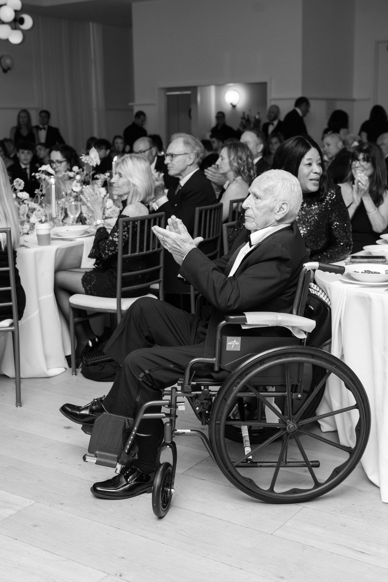 Wedding guests seated at round tables applaud during reception while an elderly gentleman watches from his wheelchair.