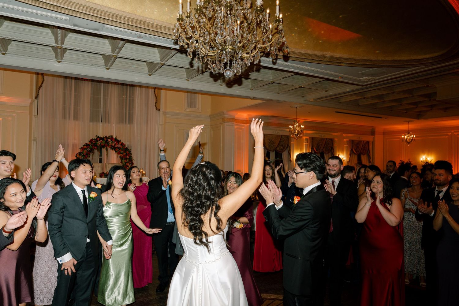 Wedding celebration with guests dancing under crystal chandeliers in an elegant ballroom.