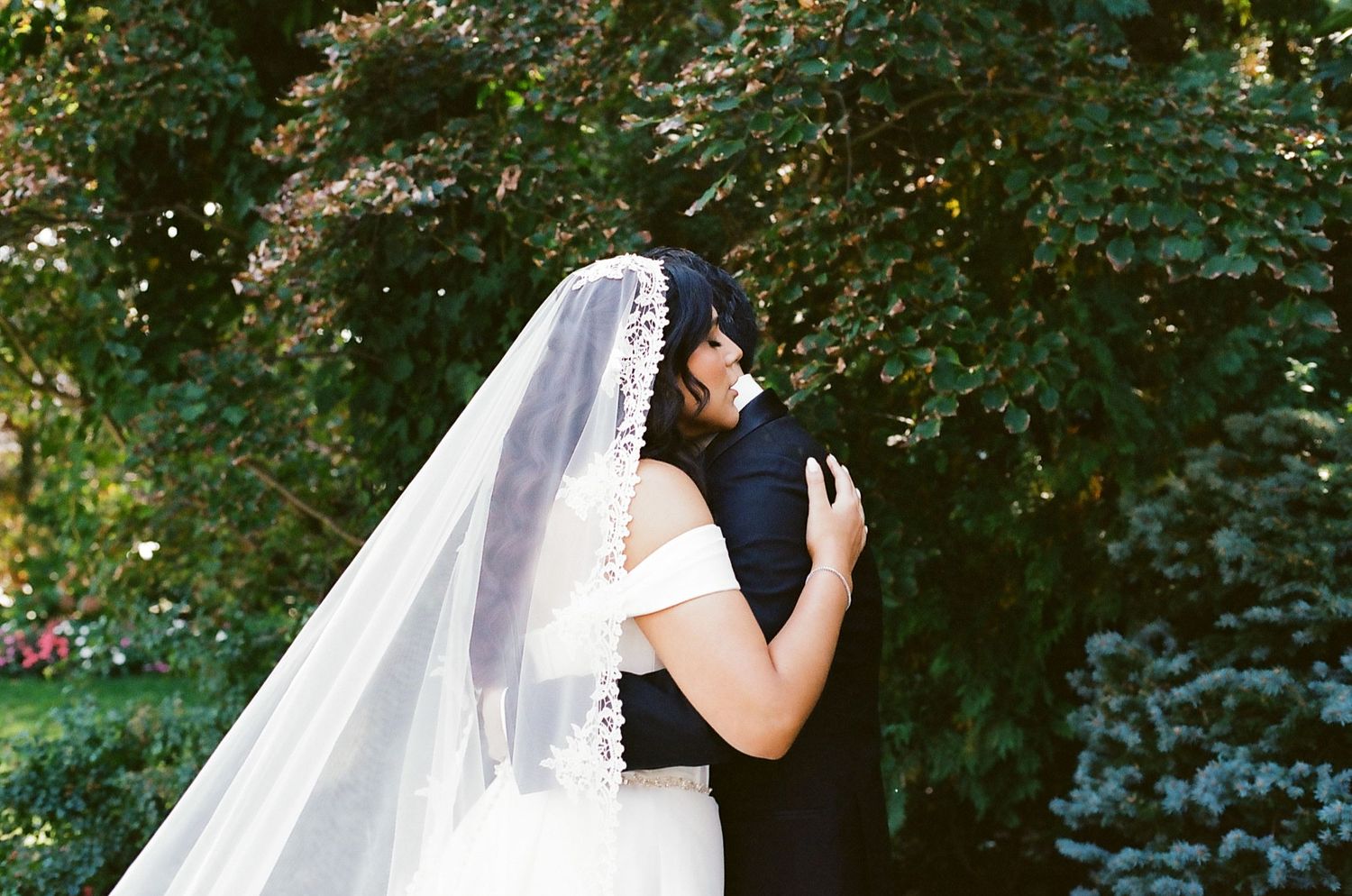 A couple in wedding attire shares a romantic embrace under lush green trees in a garden setting.