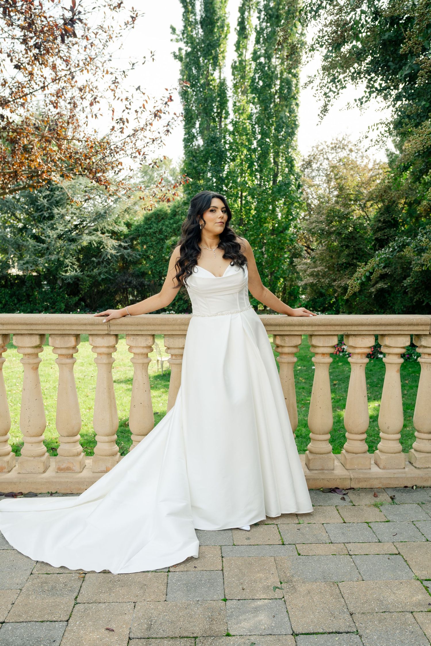 A bride in a flowing white wedding gown poses gracefully on a stone terrace with ornate balustrades and lush greenery.