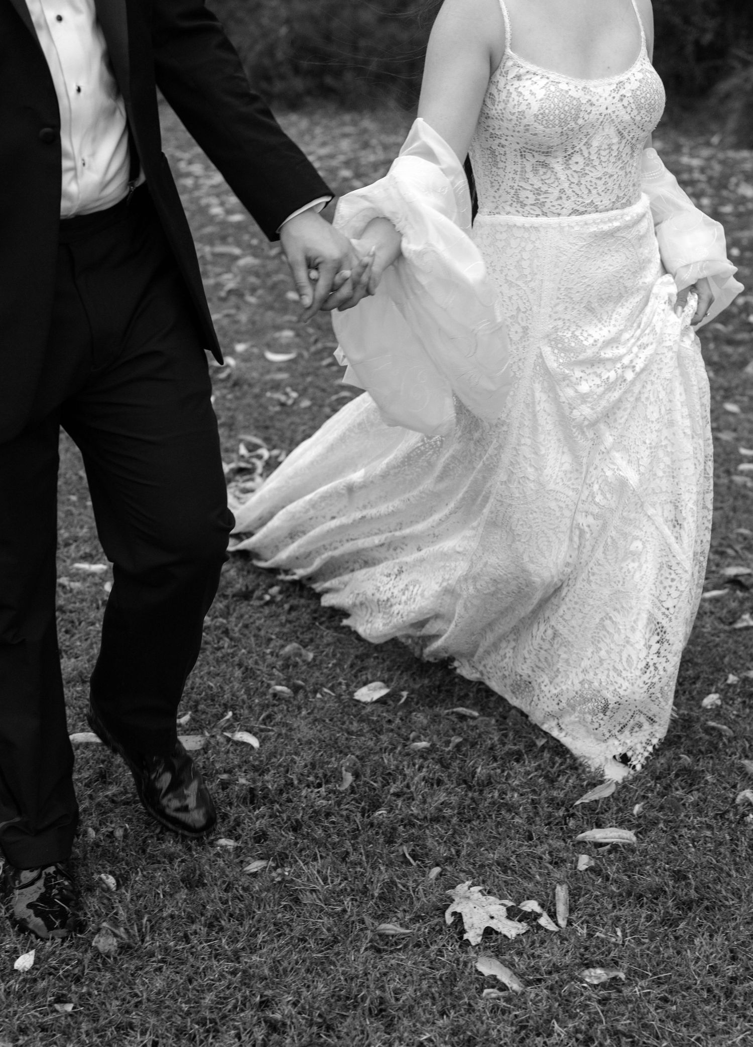 Black and white photo showing the bottom half of a couple in formal wedding attire walking together on grass.