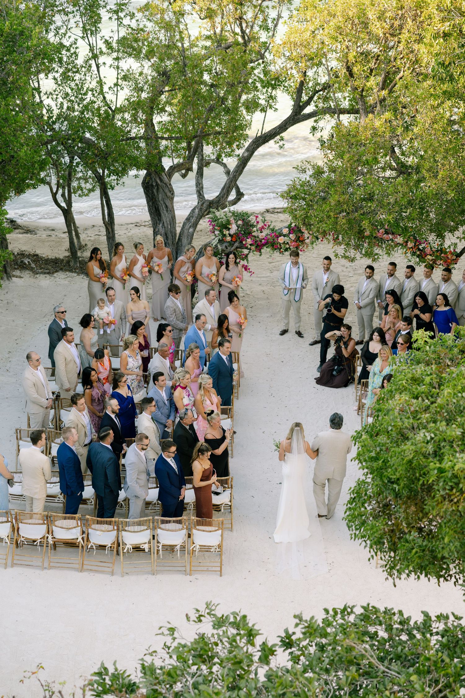 Aerial view of an outdoor wedding ceremony surrounded by trees with guests seated on white chairs.