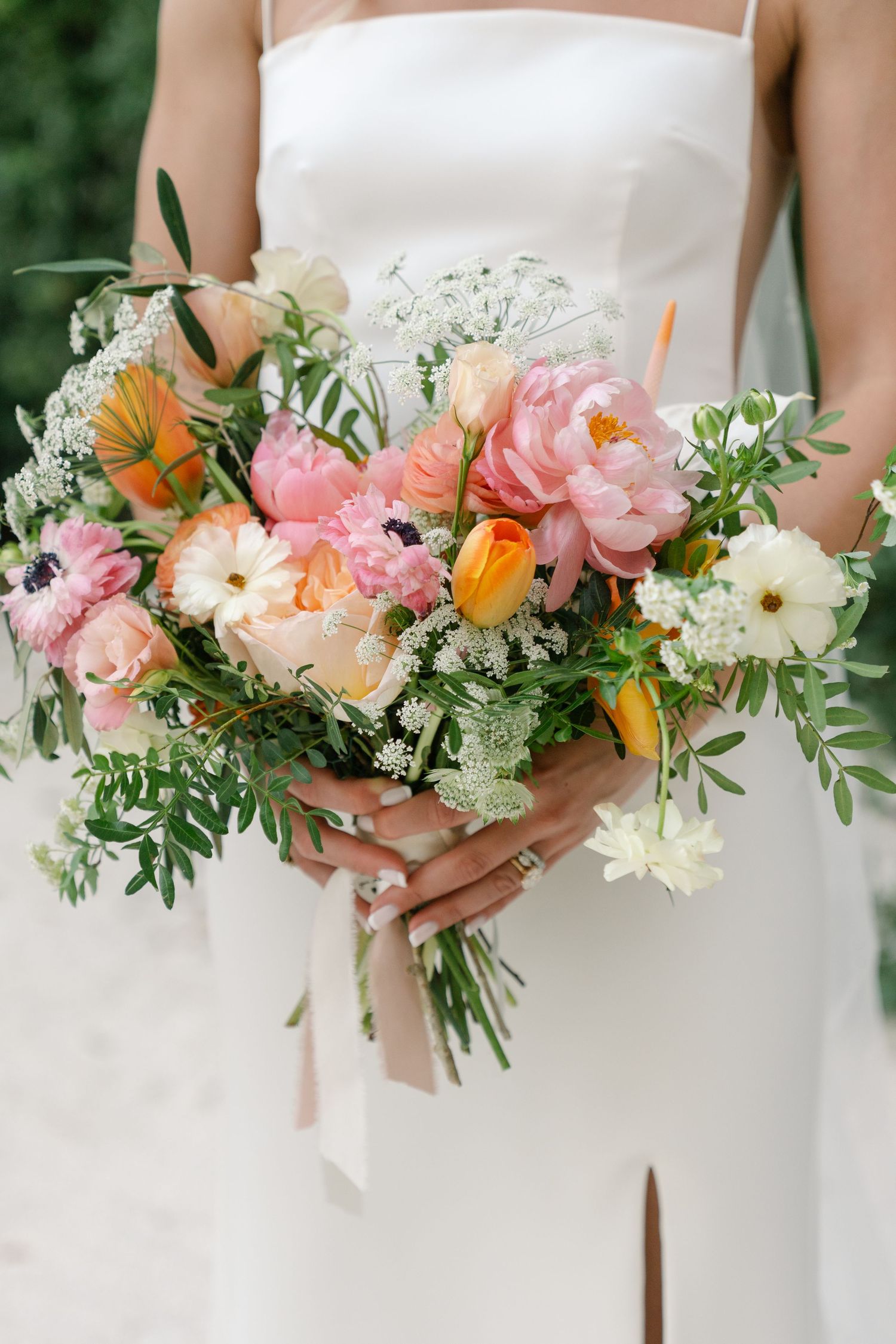 A stunning bridal bouquet featuring pink peonies, orange tulips, and white flowers with greenery against a white dress.