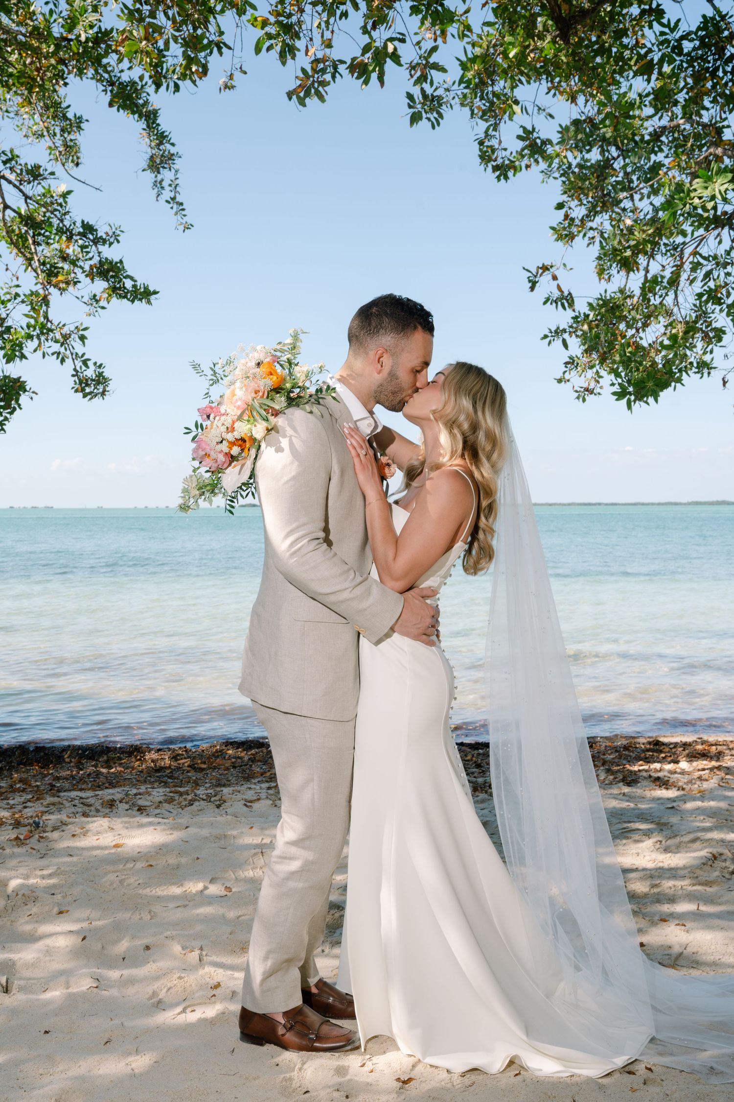Romantic beachside wedding ceremony with couple embracing under tropical trees by turquoise waters.