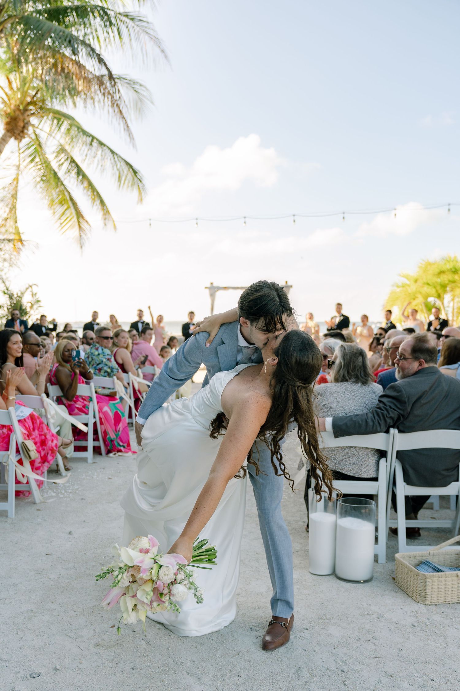 A bride and groom embrace and kiss on a sandy beach, surrounded by palm trees and a crowd of people.