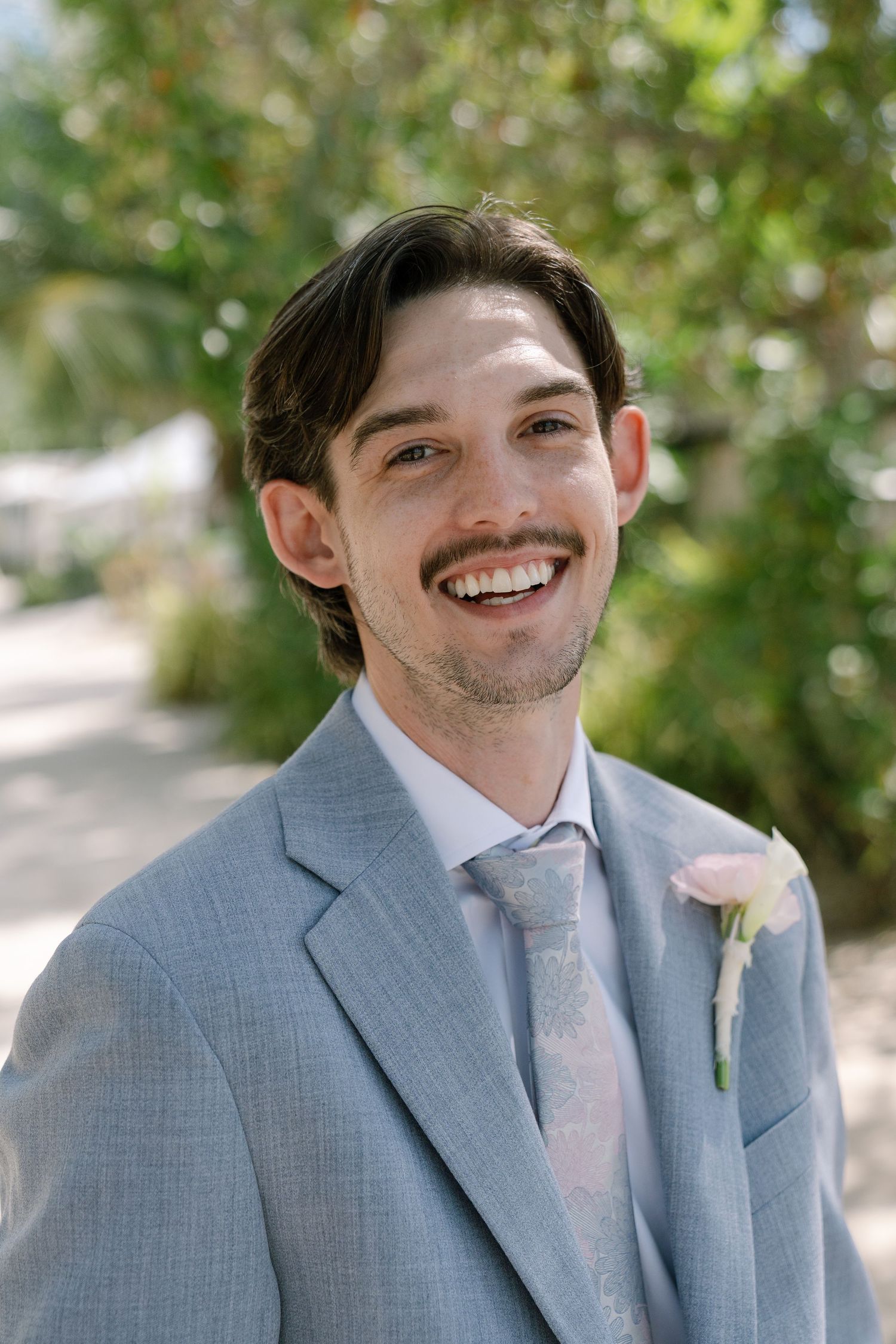A smiling man in a gray suit stands in a scenic outdoor setting.